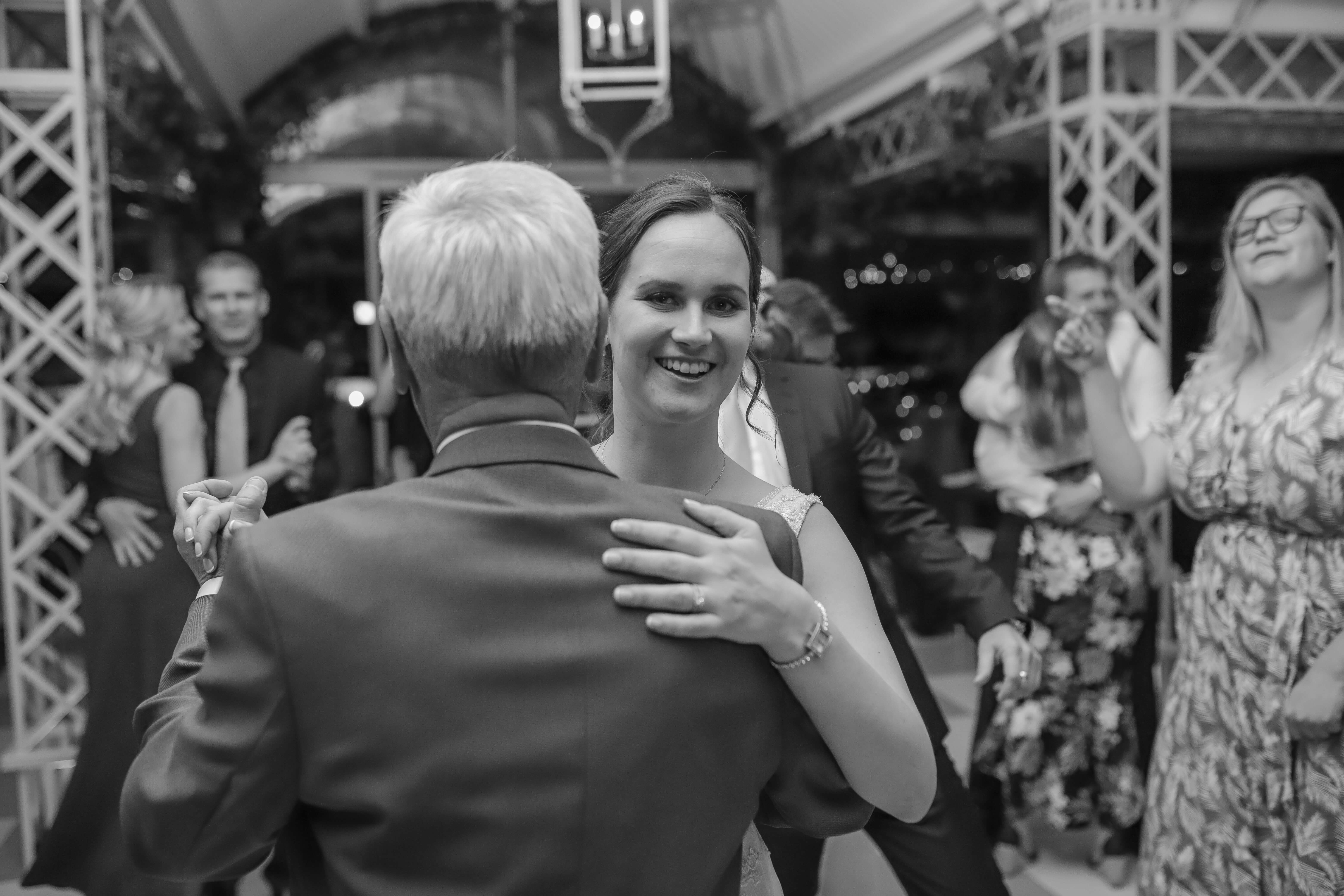 A bride smiling looking at the camera, while holding her father who is facing away.