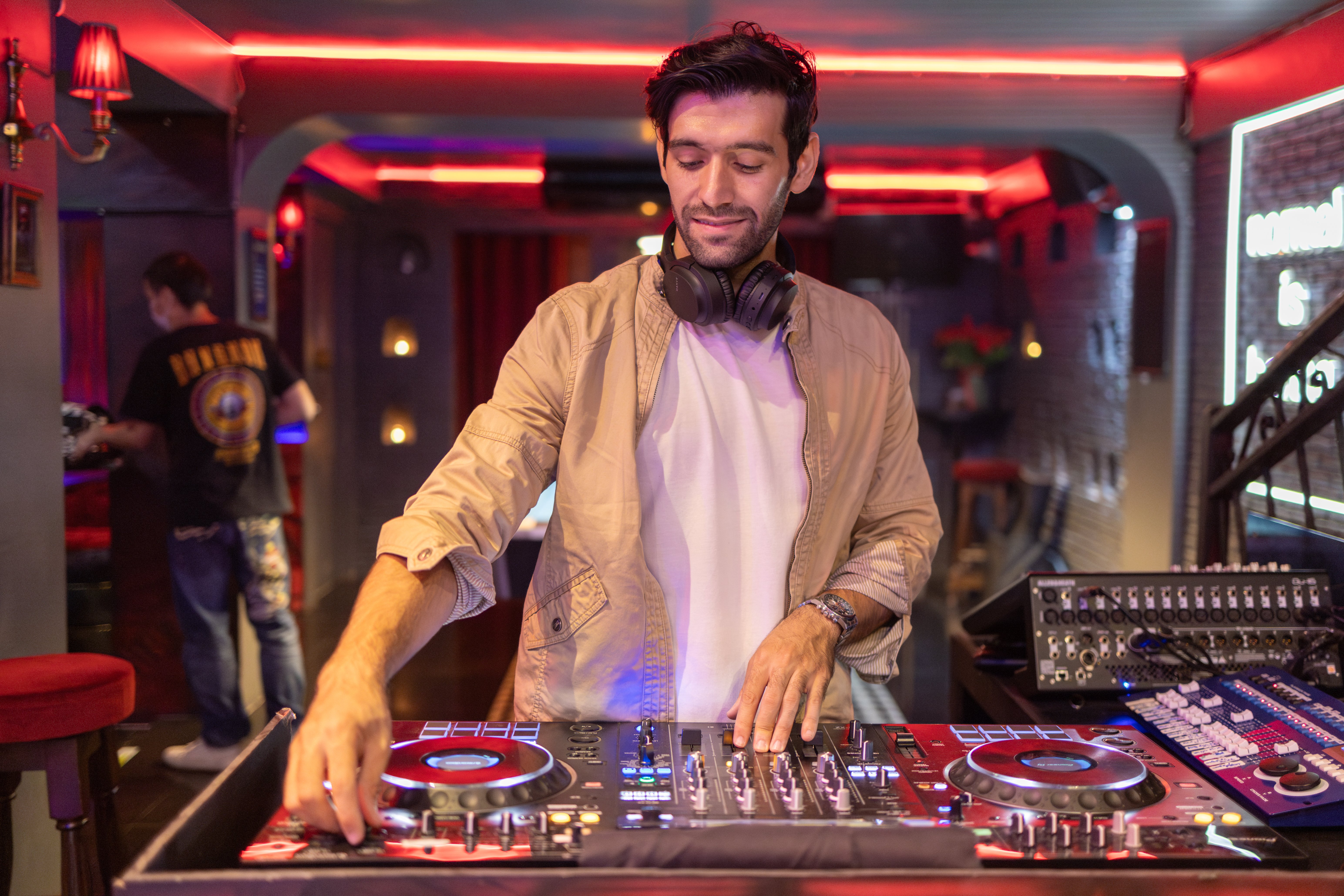 DJ with headphones round his neck mixing on a DJ controller in a red-lit bar