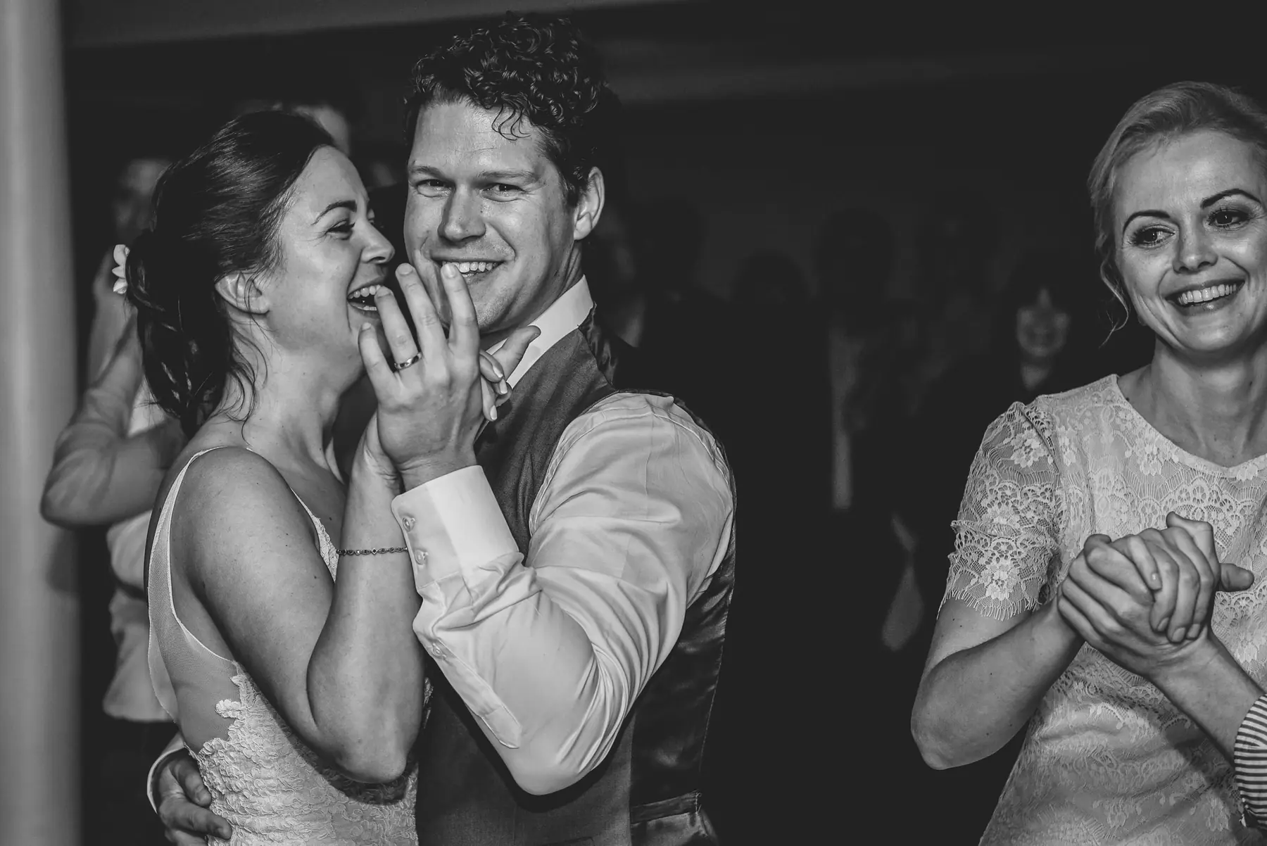 Karen and Lial laughing on the dance floor in black and white, a guest in lace smiling beside them with hands clasped