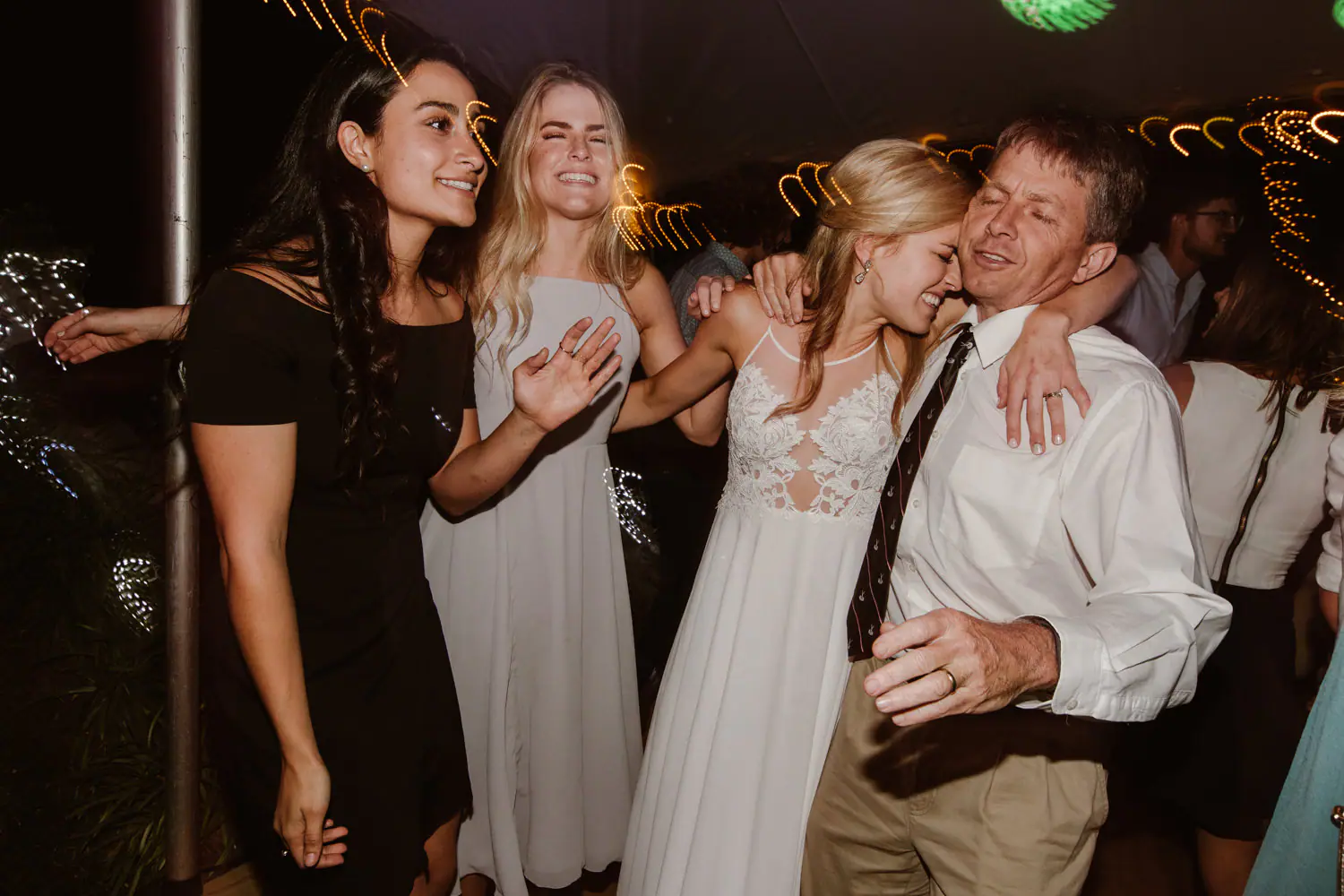 Sarah hugging an older man on the dance floor as a bridesmaid and a friend laugh beside her, fairy lights swirling overhead