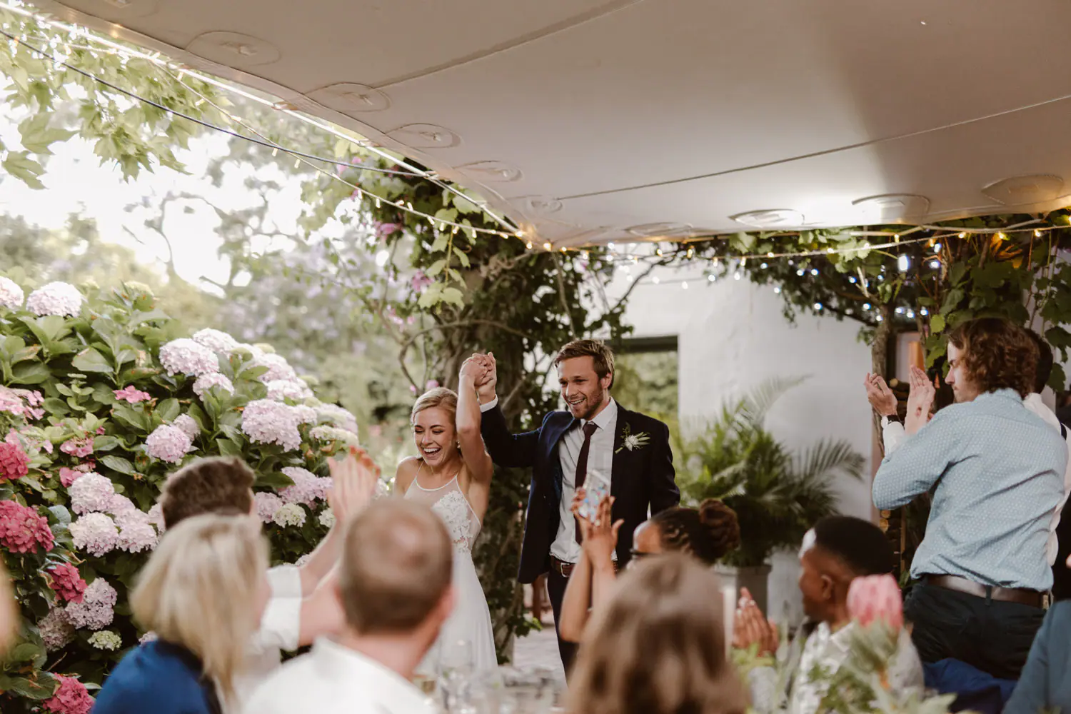 Ramsay and Sarah walking into their reception with joined hands raised as guests stand and clap beside pink hydrangeas