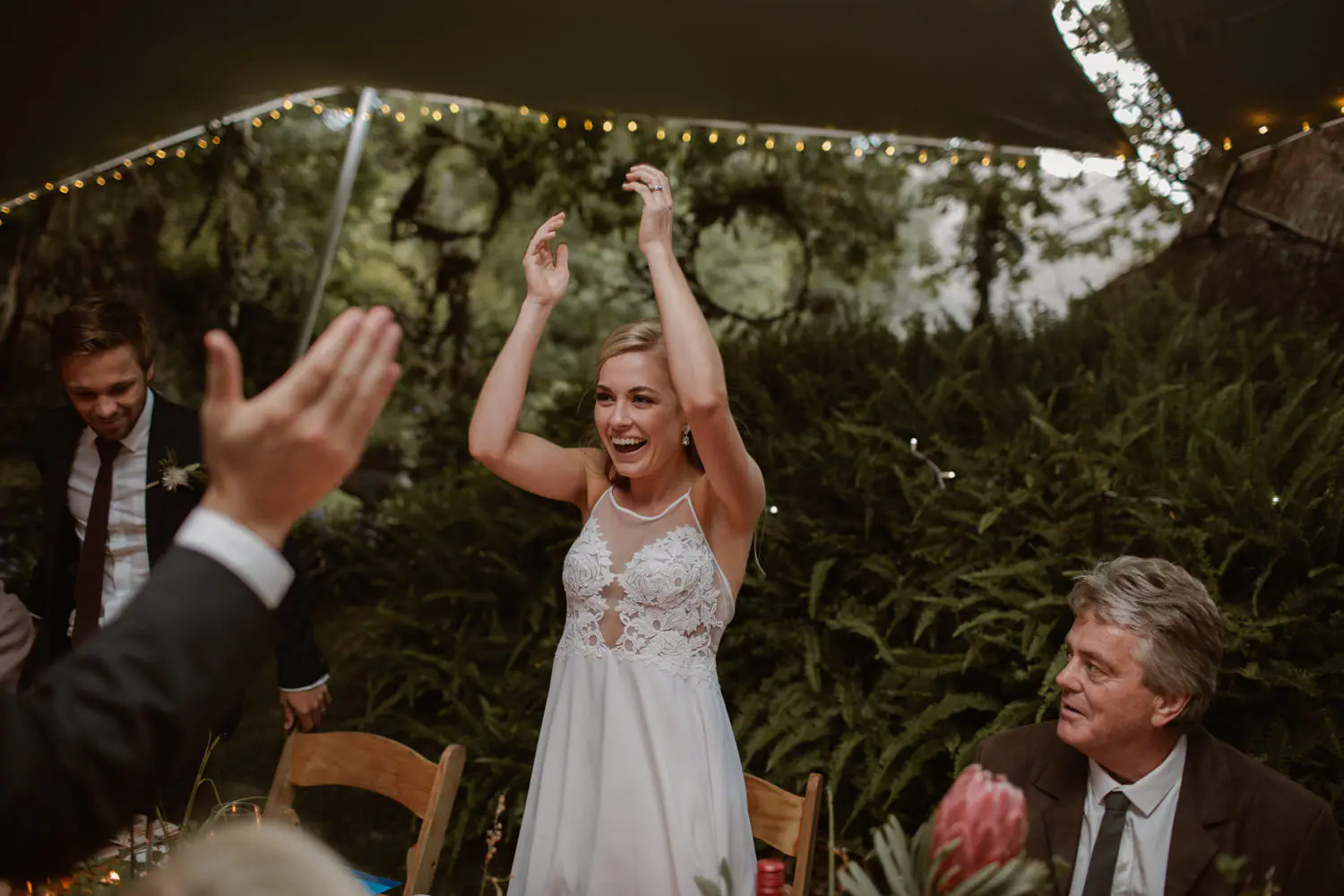 Sarah standing at the bridal table with both arms raised and laughing, her father seated beside a pink king protea