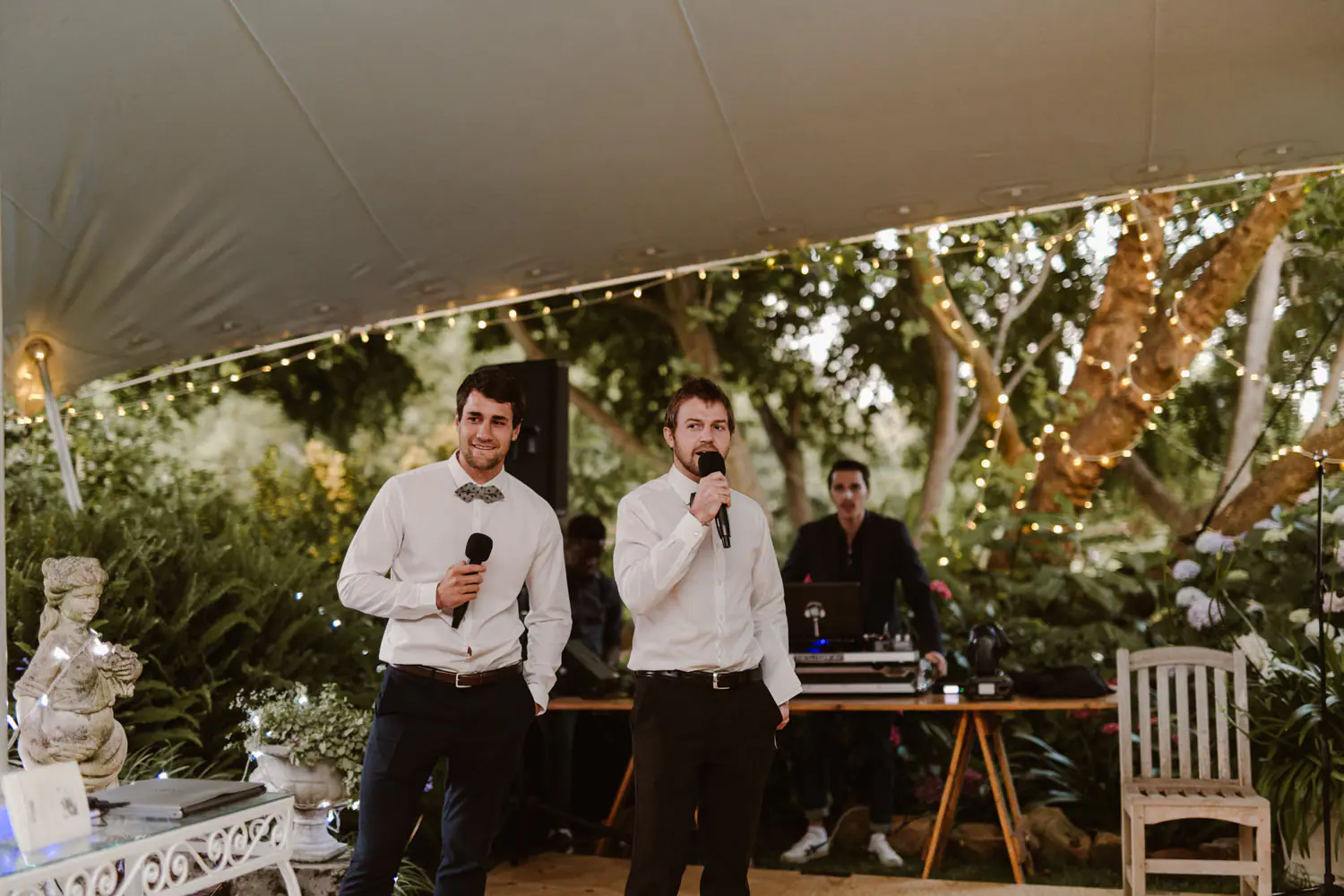 Two men speaking on wireless microphones as the DJ waits at his laptop and mixer behind them, festoon lights in the trees