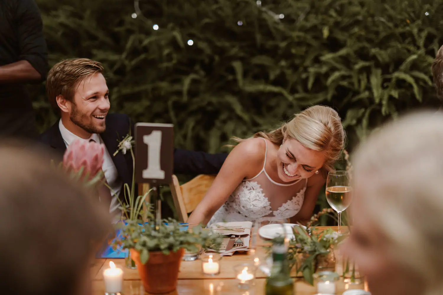 Sarah and Ramsay laughing at table number 1 during the speeches, with a pink king protea, potted greenery and tea lights