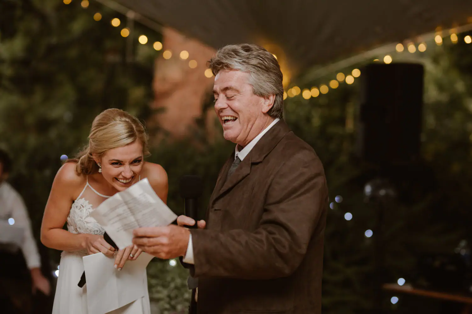 Sarah and her father laughing through a joint speech, both holding microphones and pages, a speaker beside them
