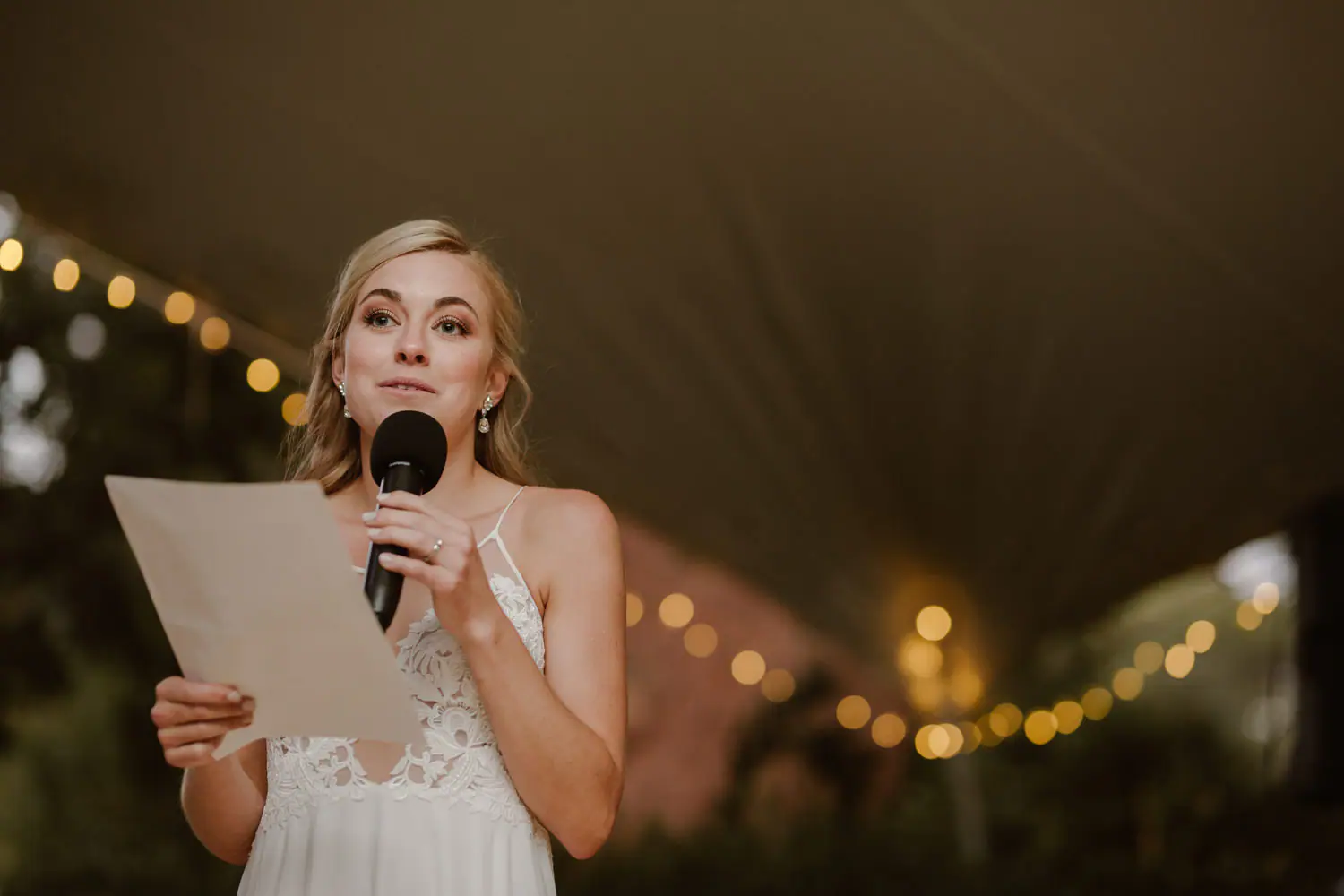 Sarah giving her speech into a handheld microphone, page in hand, festoon bulbs blurred beneath the marquee roof