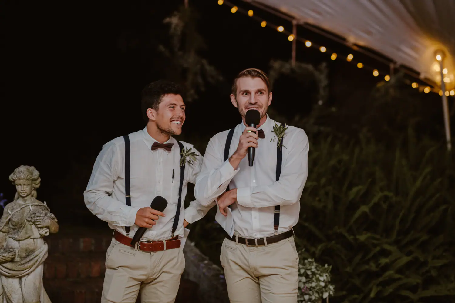 Two groomsmen in braces and burgundy bow ties grinning through a speech, microphones in hand, beside a stone garden statue