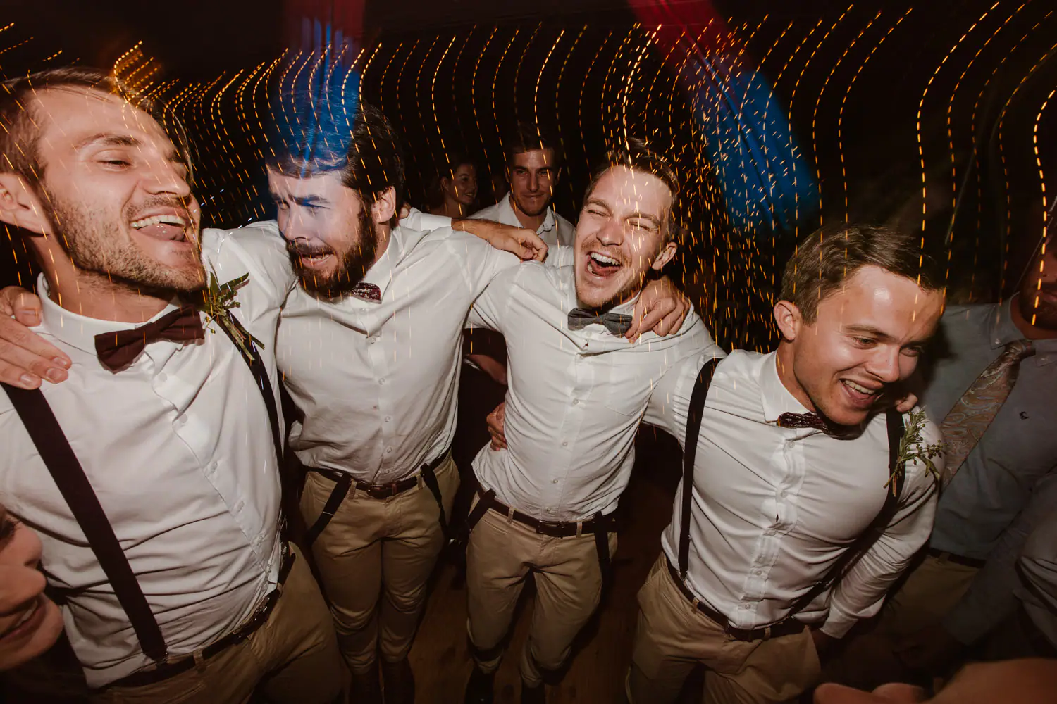 Four groomsmen in braces and bow ties jumping arm in arm and shouting, fairy lights streaking behind them