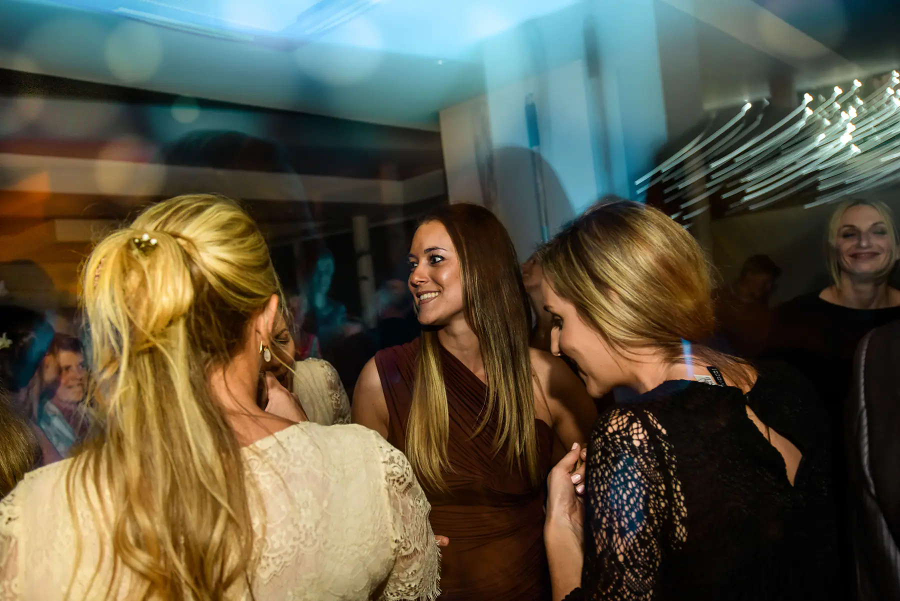 Three women in brown, cream lace and black lace chatting on the dance floor under cool blue light in Plettenberg Bay