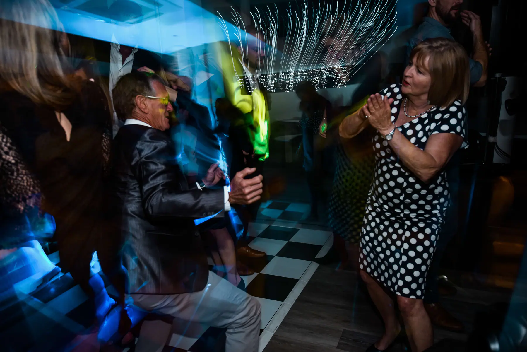 Man in tinted glasses crouching mid-dance as a woman in a polka dot dress claps, chandelier light streaking above