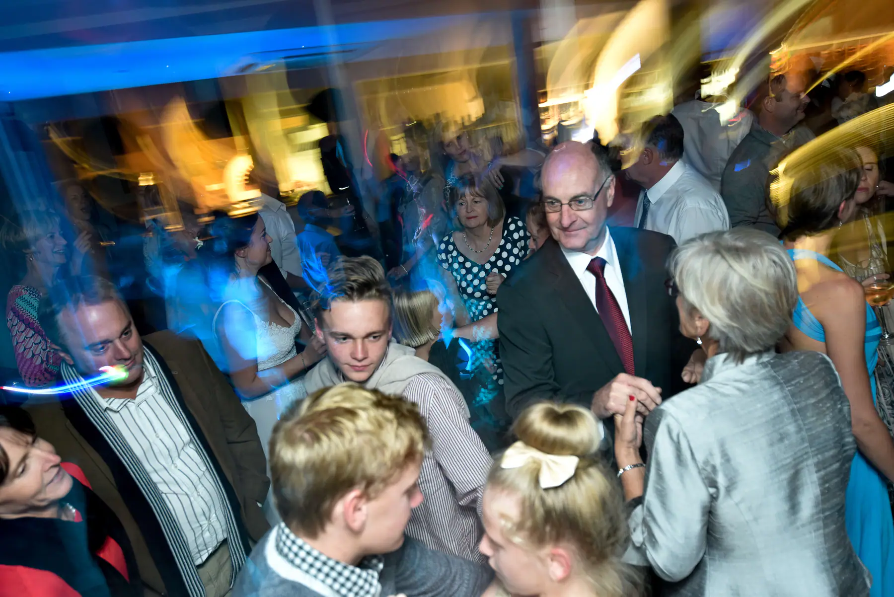 Man in a red tie dancing hand in hand with a woman in a silver jacket amid a crowd of guests of all ages