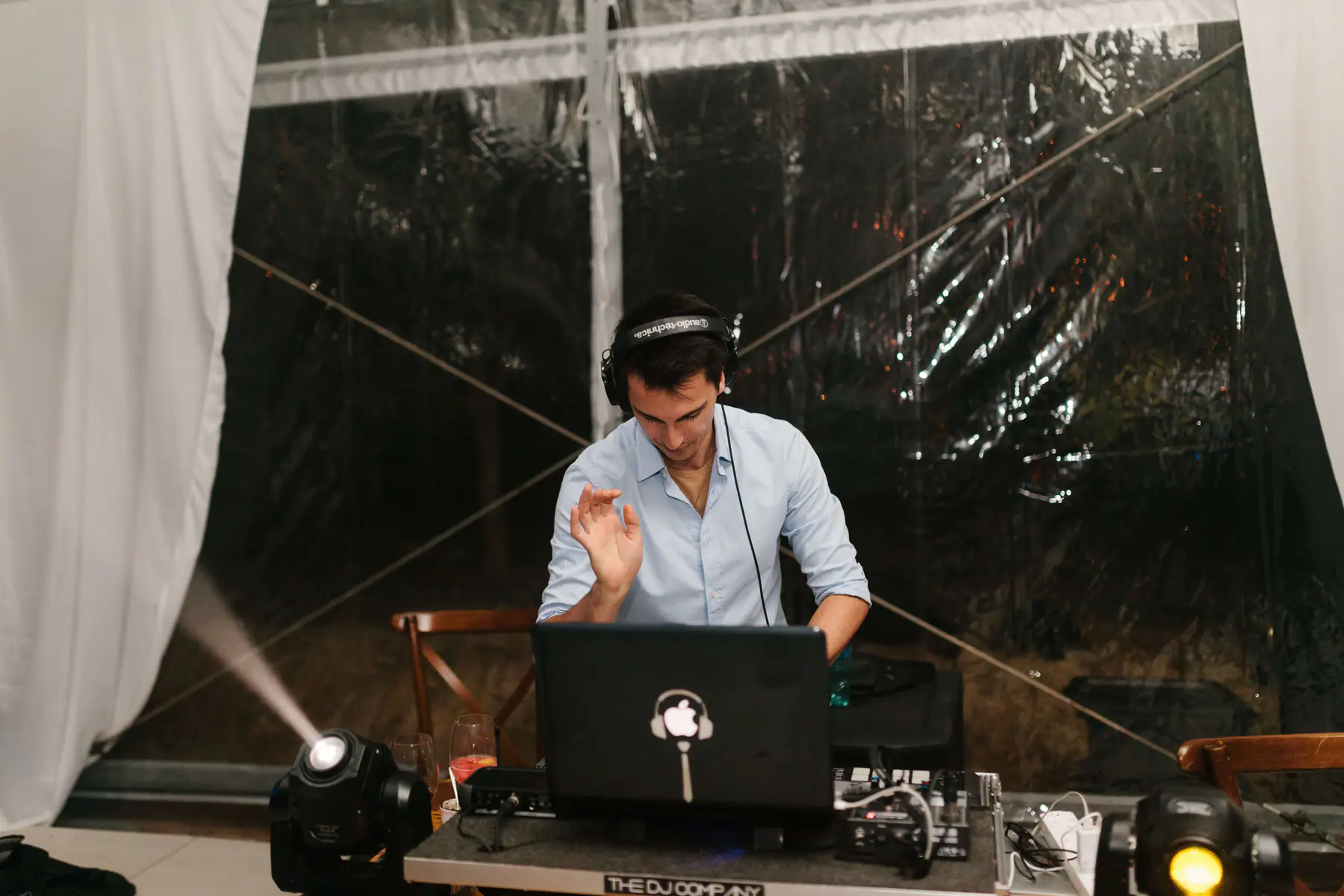 The DJ Company's DJ in headphones mixing on a laptop and controller between two moving head lights in a clear marquee