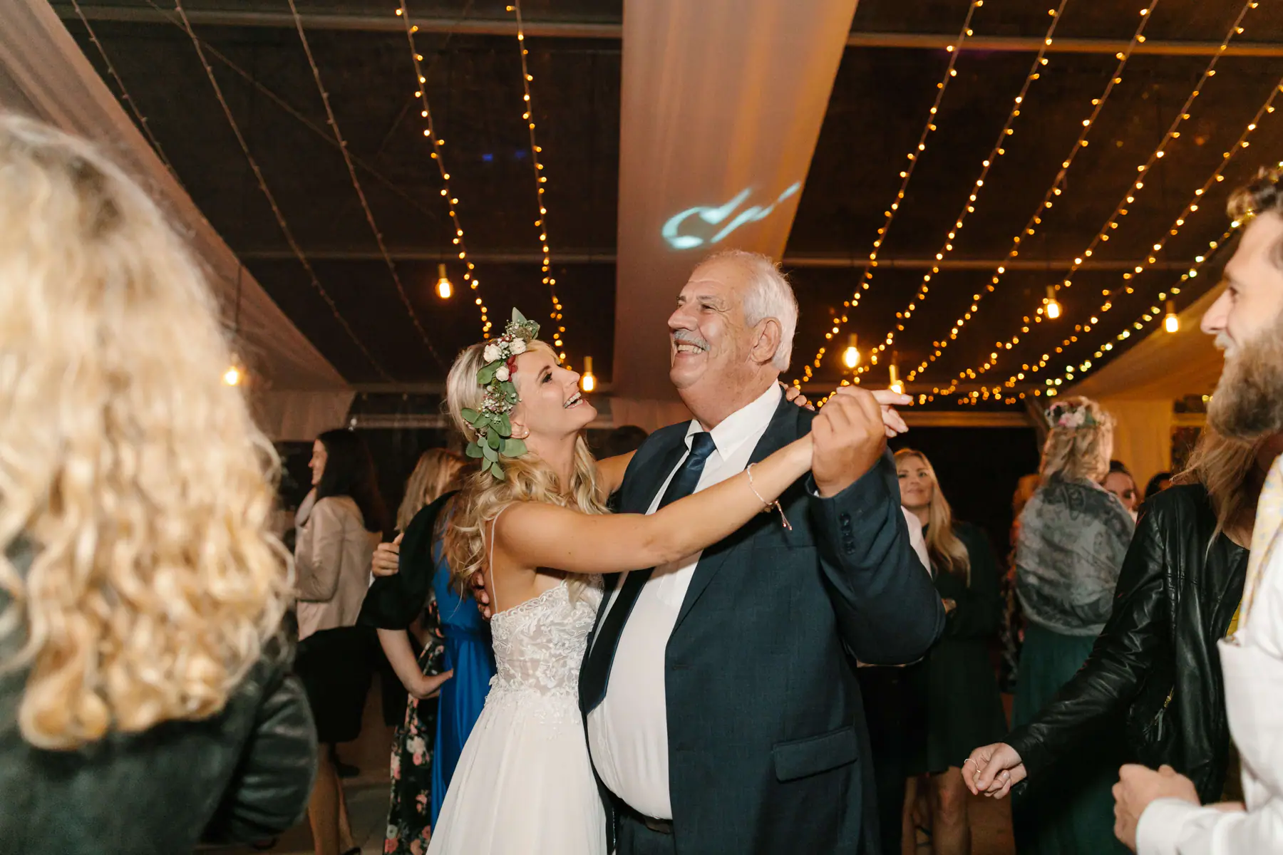Louisa in a flower crown laughing as she dances with a grey-haired man in a navy suit under strings of fairy lights