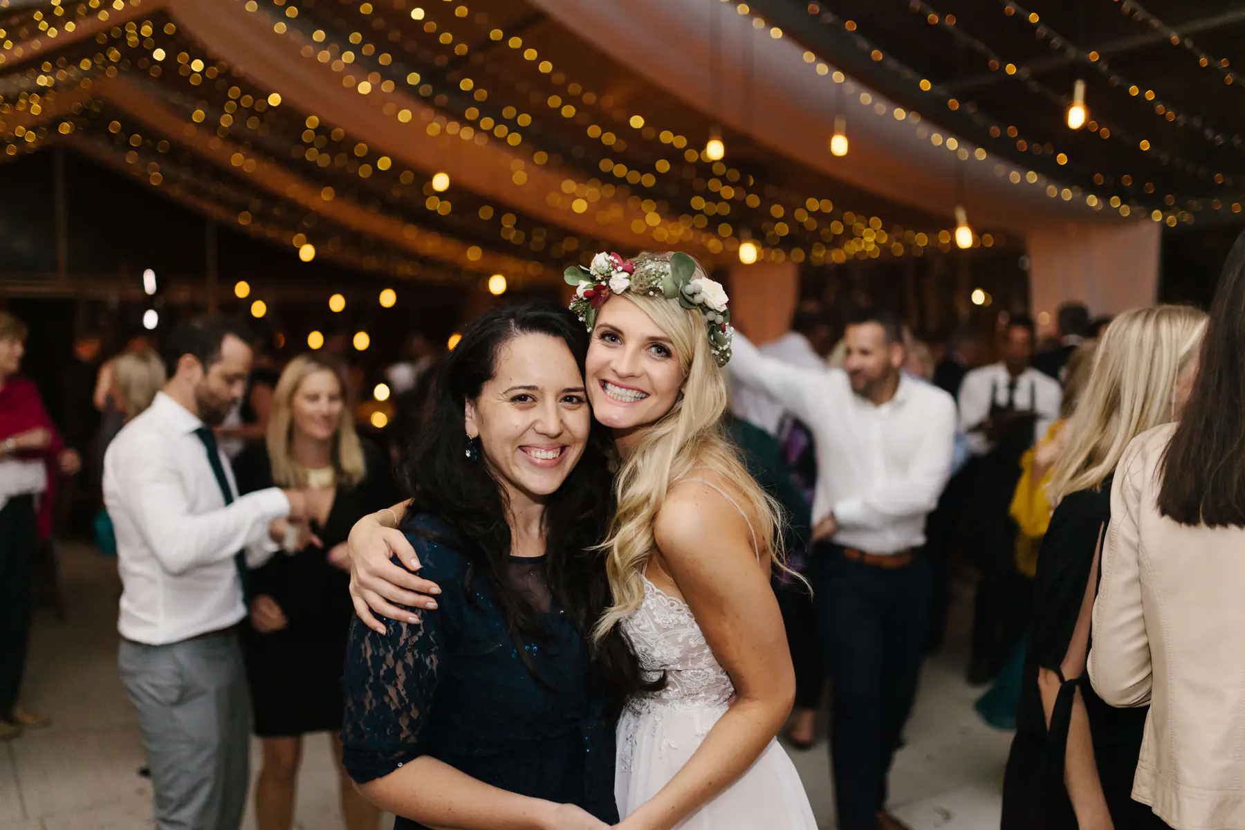 Louisa in a flower crown hugging a dark-haired friend in navy lace under a canopy of fairy lights