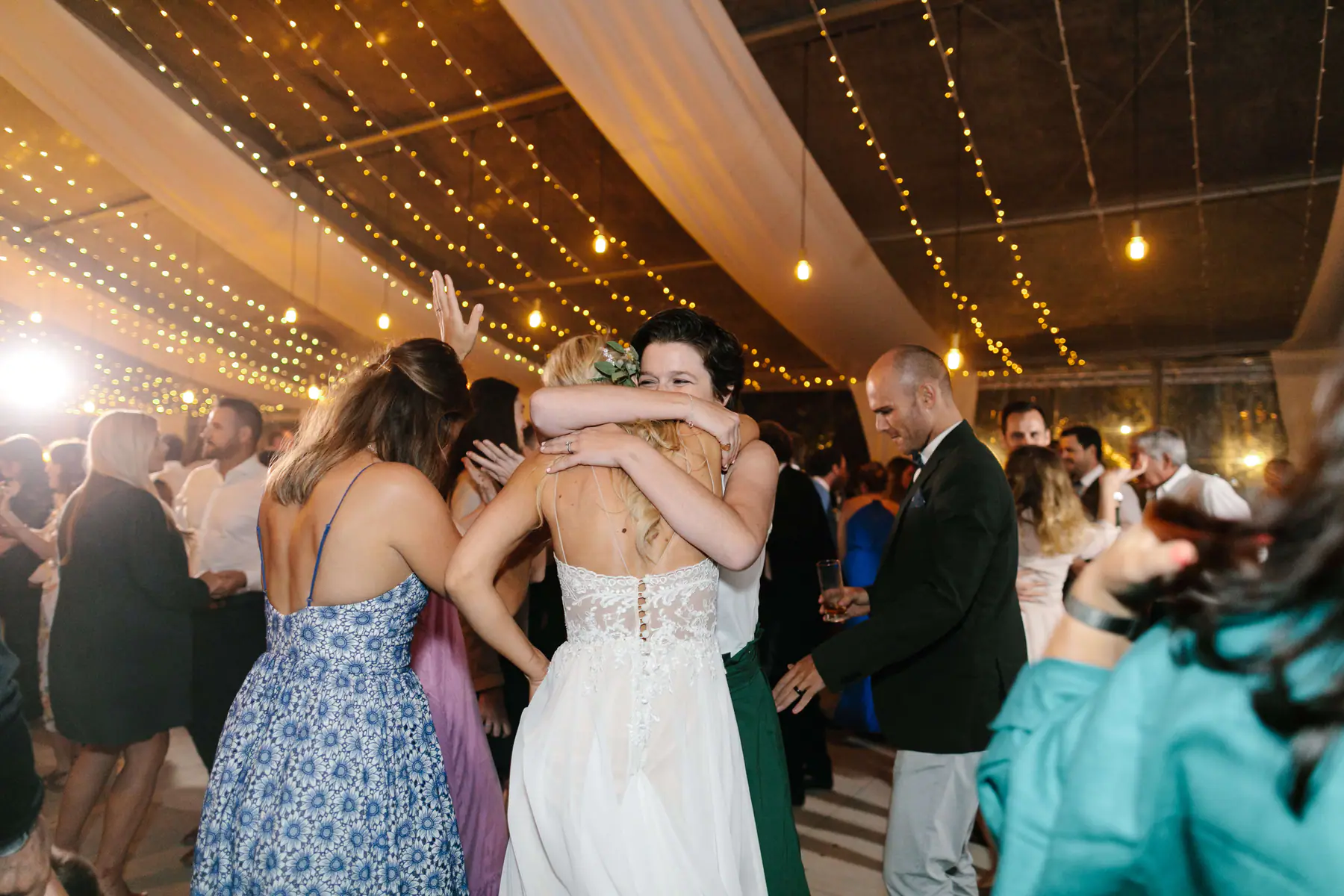 Louisa hugging a short-haired friend, the lace back of her wedding dress to the camera, fairy lights overhead