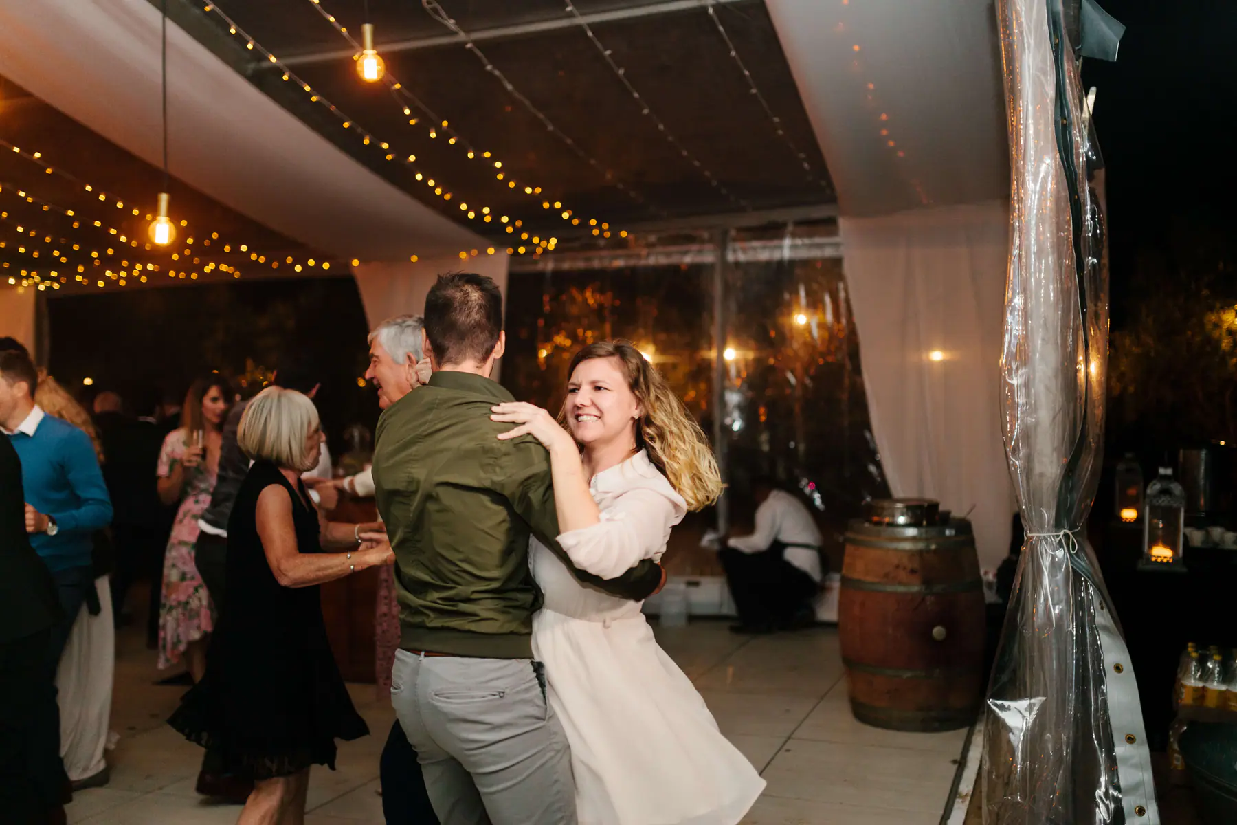 Guest in a white dress dancing with a man in an olive bomber jacket beside a wine barrel at the marquee's edge
