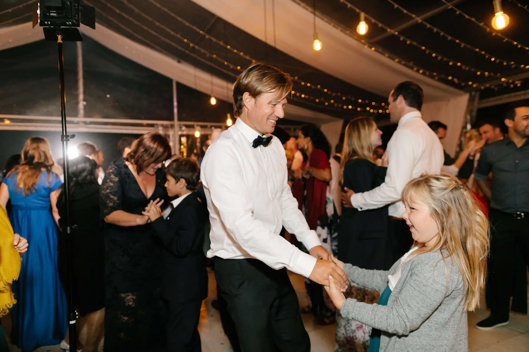 Man in a white shirt and bow tie dancing hand in hand with a young blonde guest beside a lighting stand