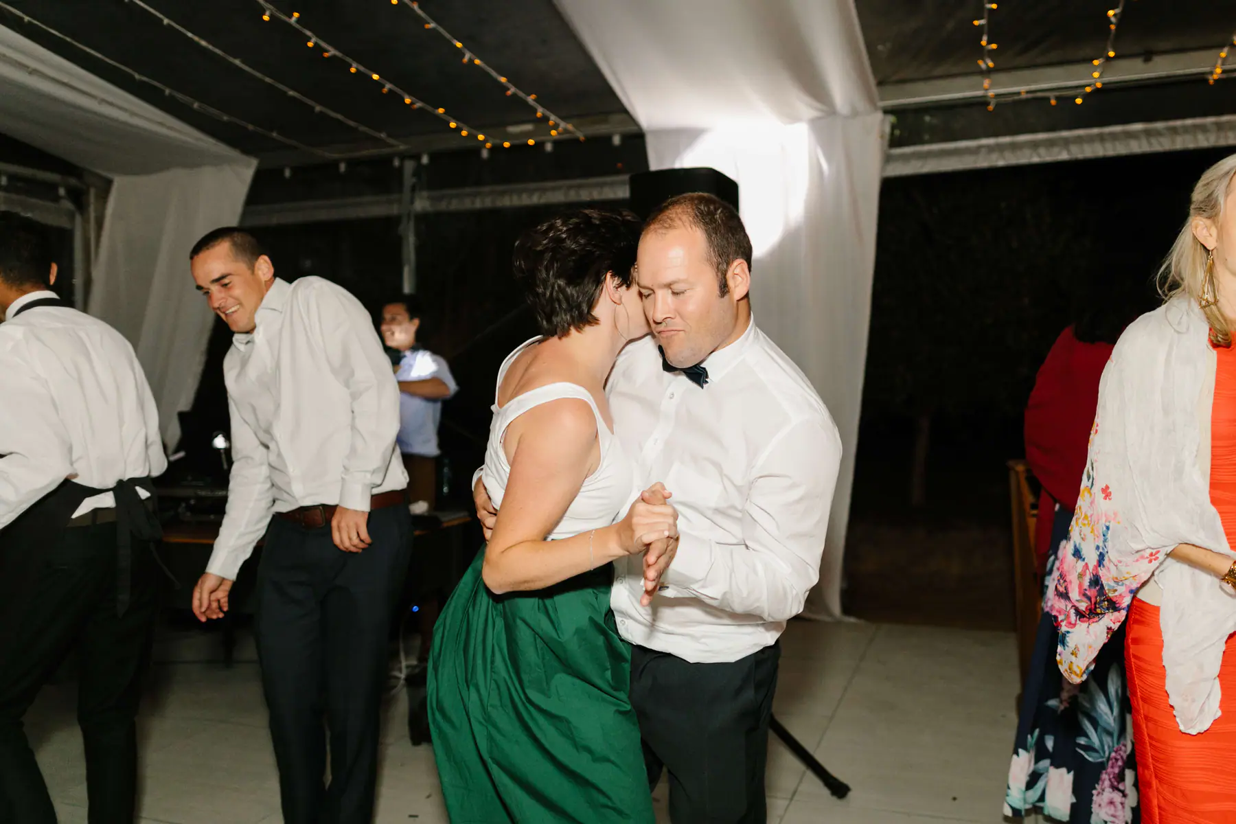 A woman in a green skirt and a man in a bow tie dance cheek to cheek as the DJ smiles from the booth behind them