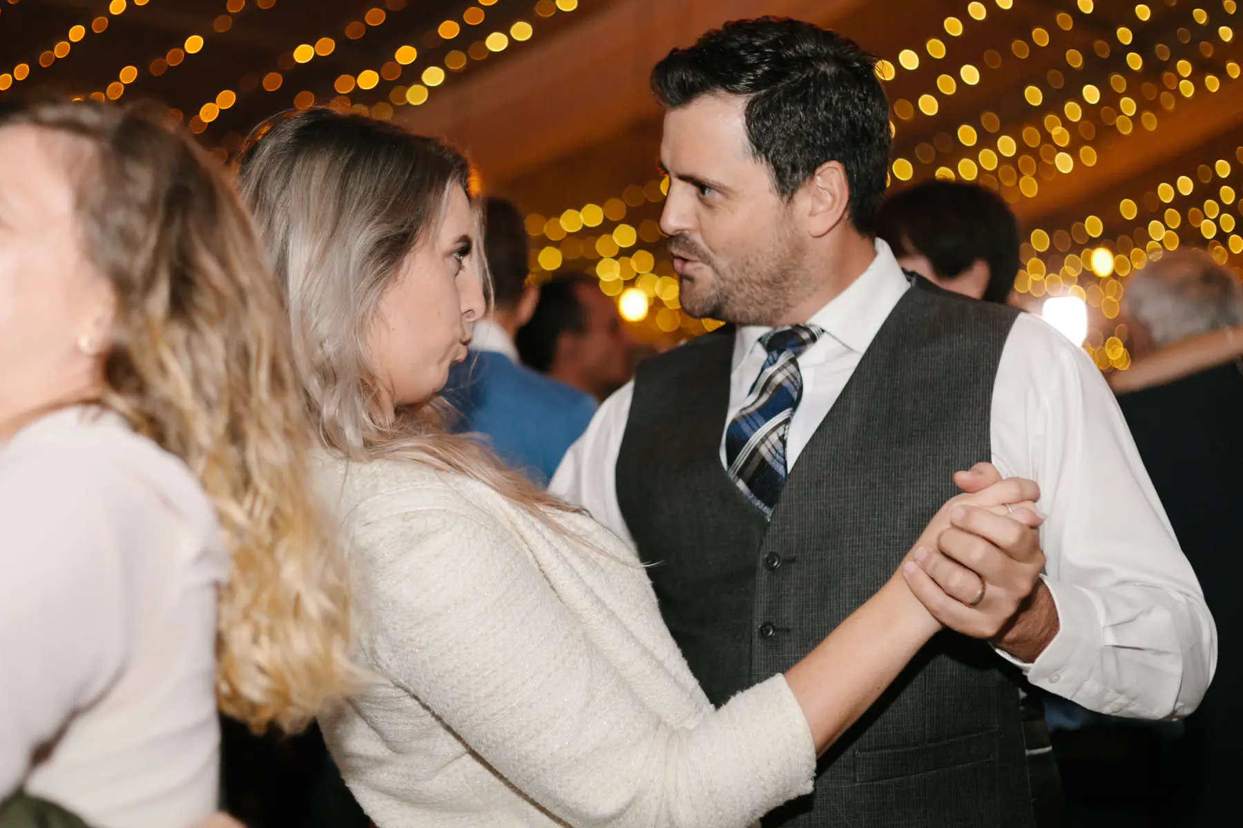 Man in a grey waistcoat and tartan tie ballroom dancing with a woman in a cream jacket under glowing fairy lights