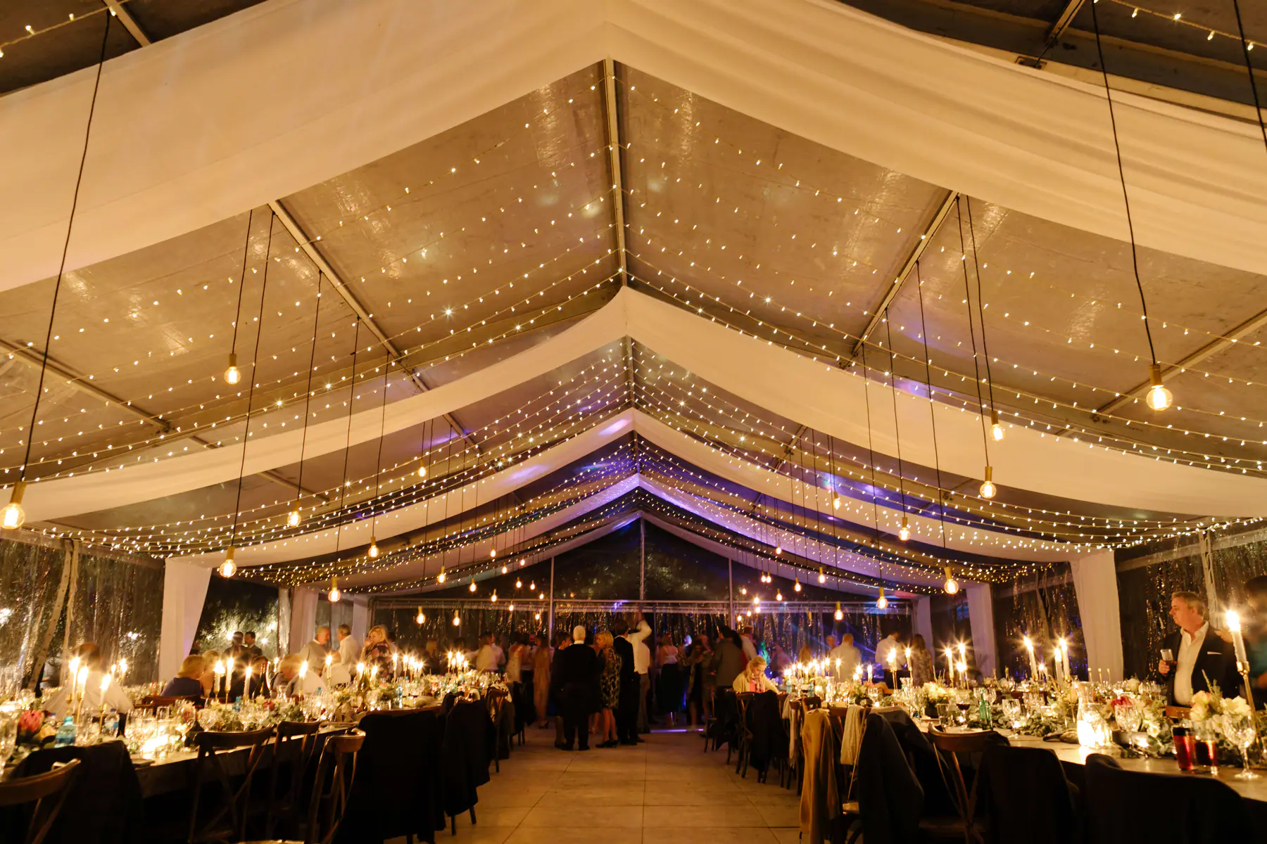 Long candlelit tables under draped fairy light canopies at the marquee reception of a Stellenbosch olive farm wedding