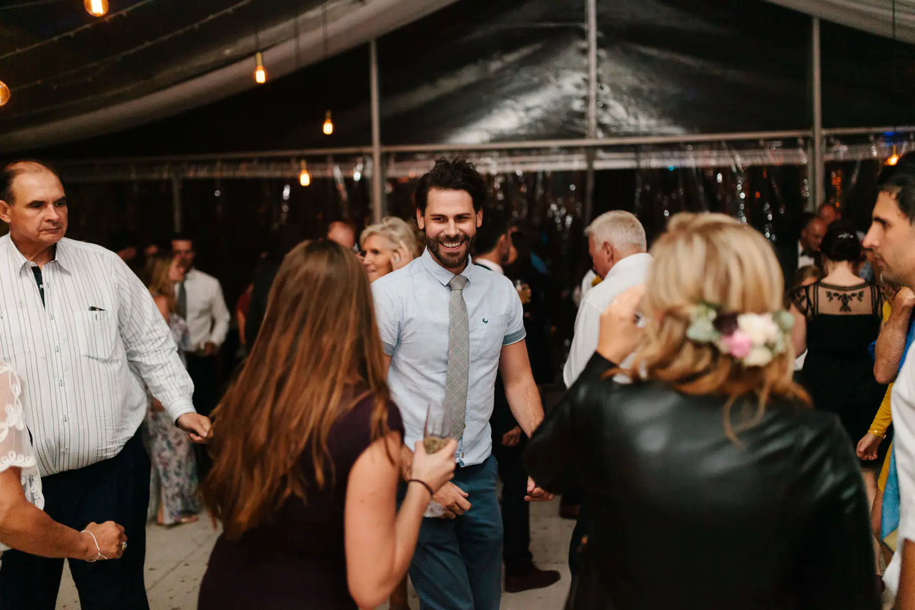 Bearded guest in a light blue shirt and grey tie grinning with a champagne glass among dancing guests