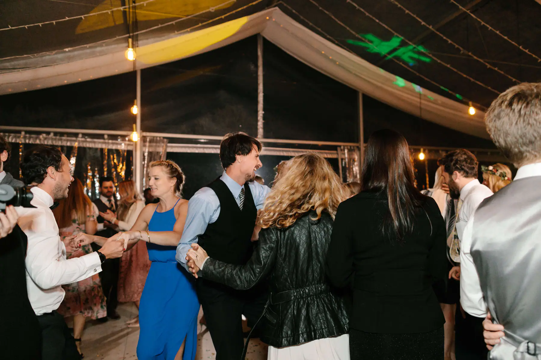 Guests linking hands in a circle dance under green and yellow gobo projections in the olive farm marquee