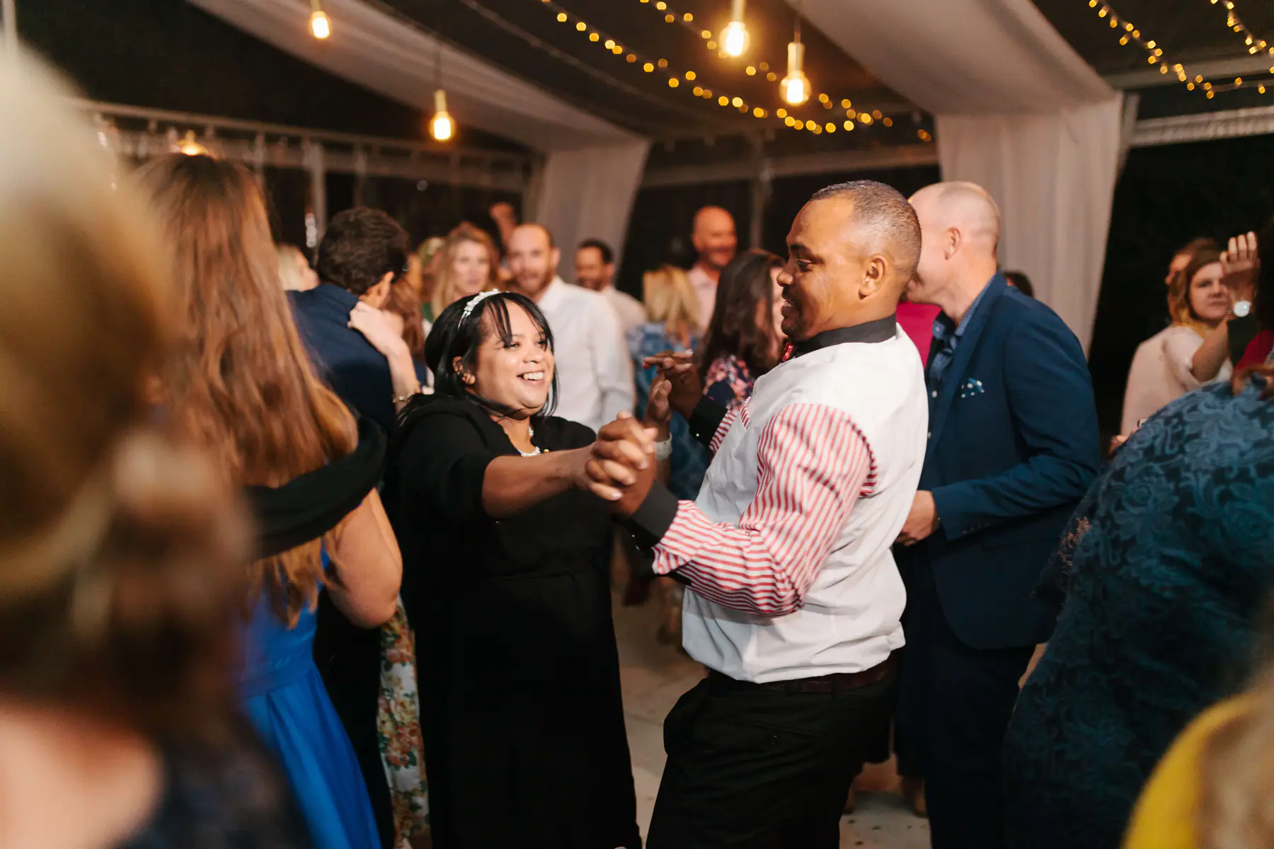 Man in a white waistcoat and striped shirt dancing hand in hand with a laughing woman in black on a busy dance floor
