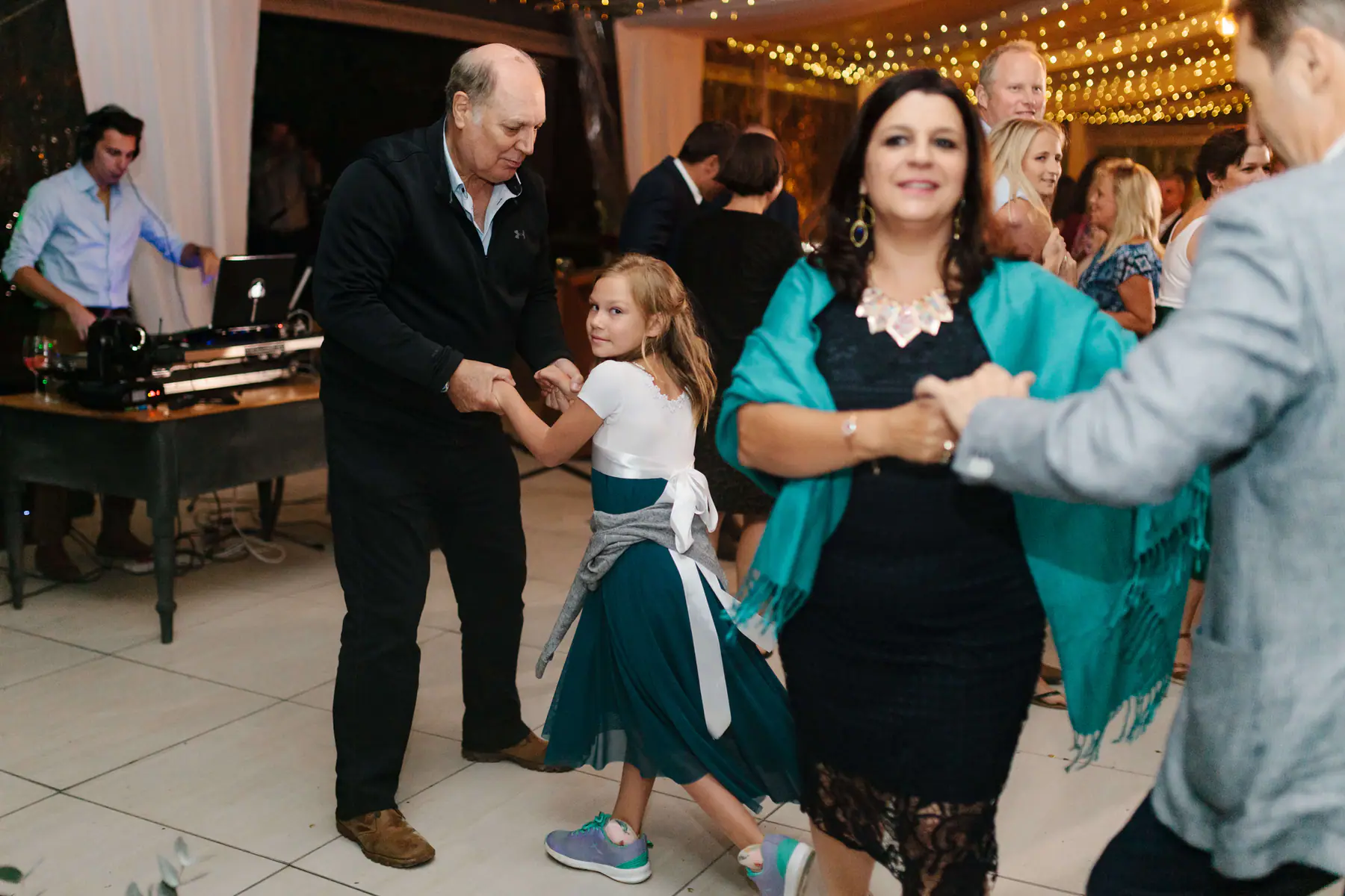 An older man dancing with a young guest in a teal dress while the DJ plays at the laptop booth behind them