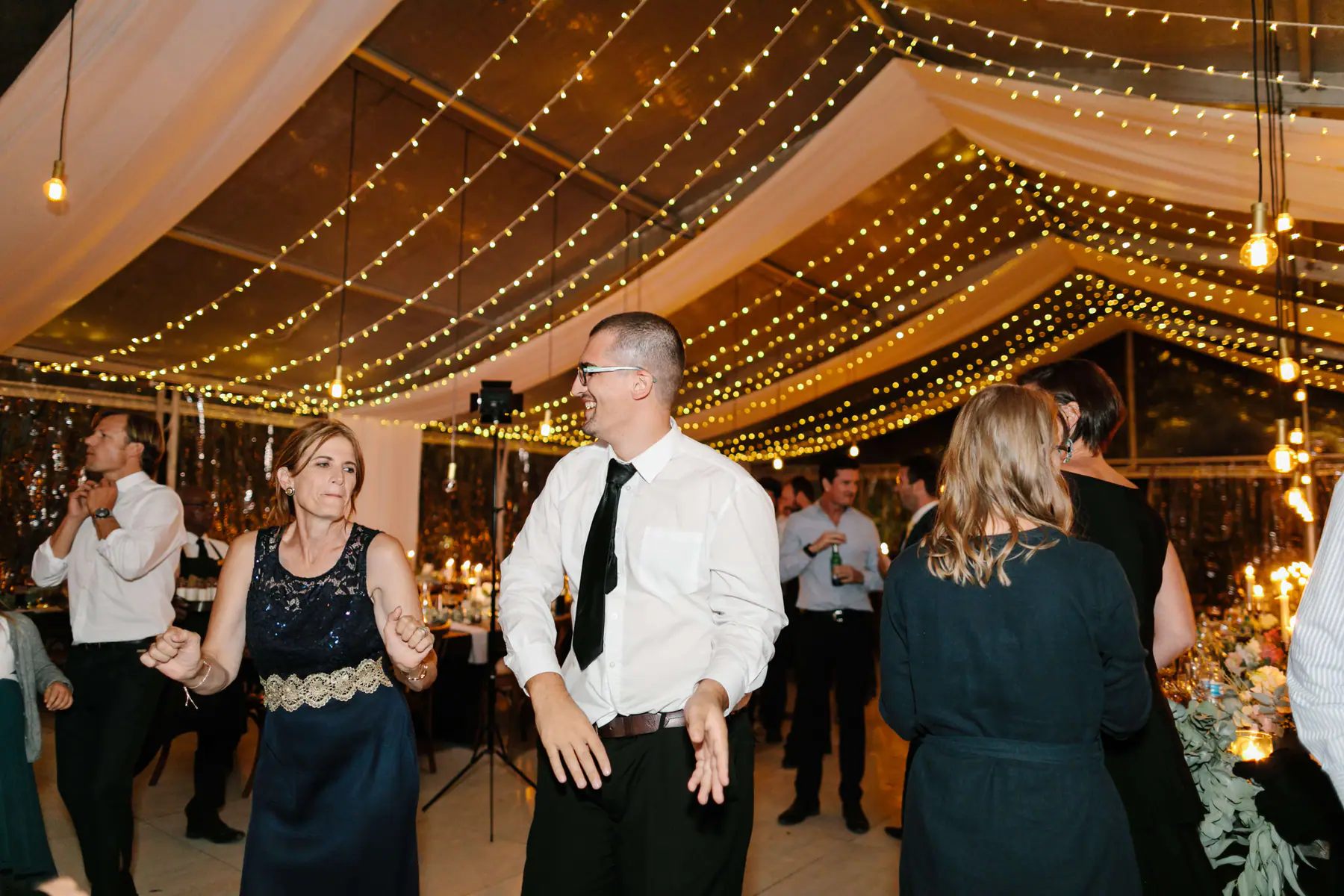 Laughing man in glasses and a black tie dancing with a woman in navy lace, candlelit tables behind them