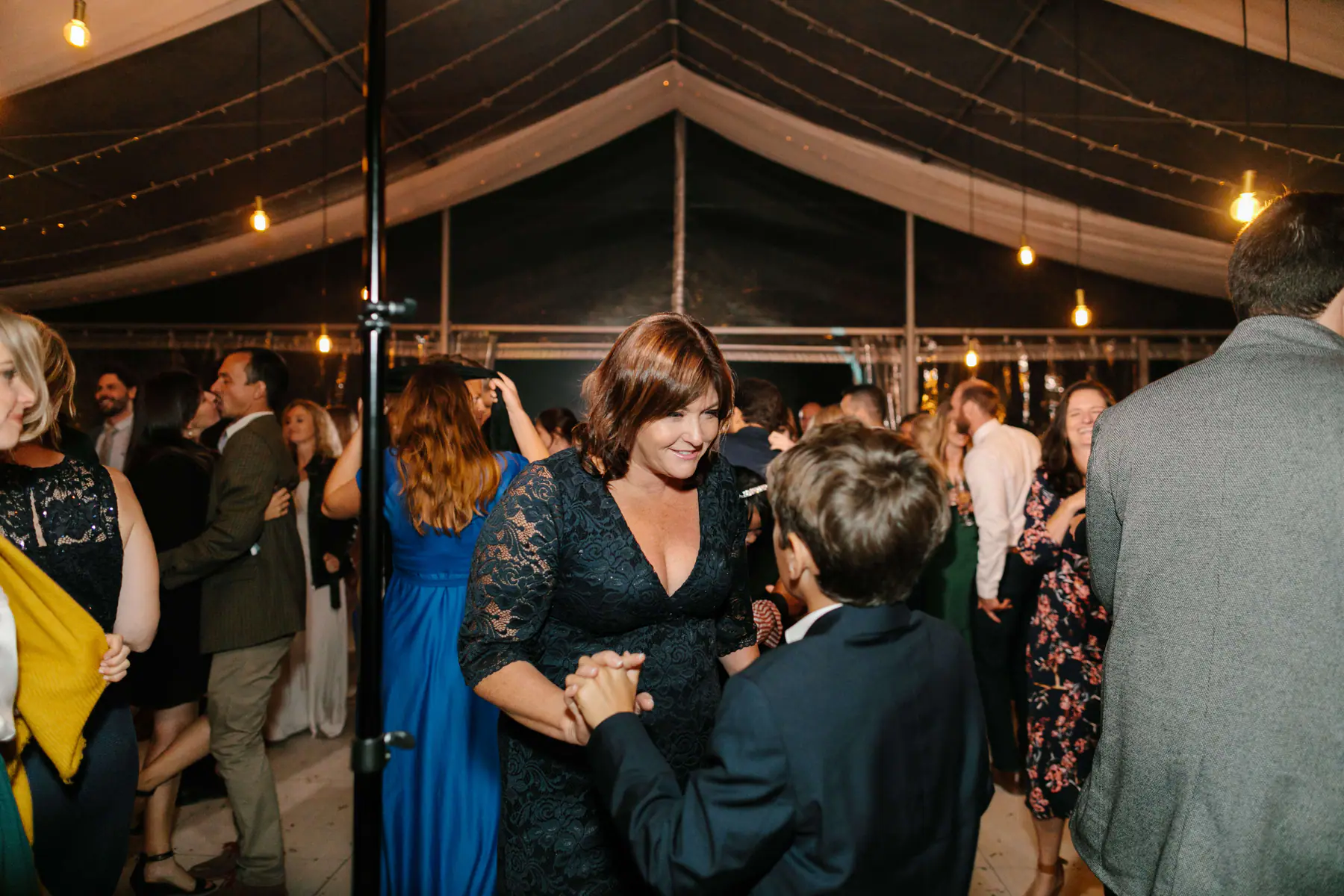 Woman in green lace dancing hand in hand with a young guest in a navy suit on a busy dance floor