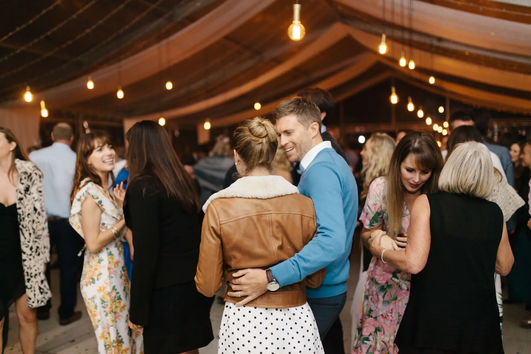 Man in a light blue jumper hugging a woman in a tan leather jacket on the dance floor under Edison bulbs
