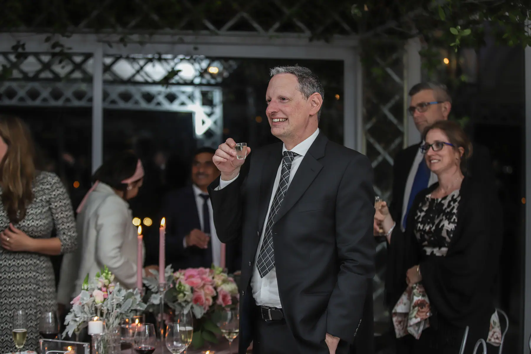 Guest in a dark suit raising a shot glass for a toast, pink roses and taper candles on the table beside him