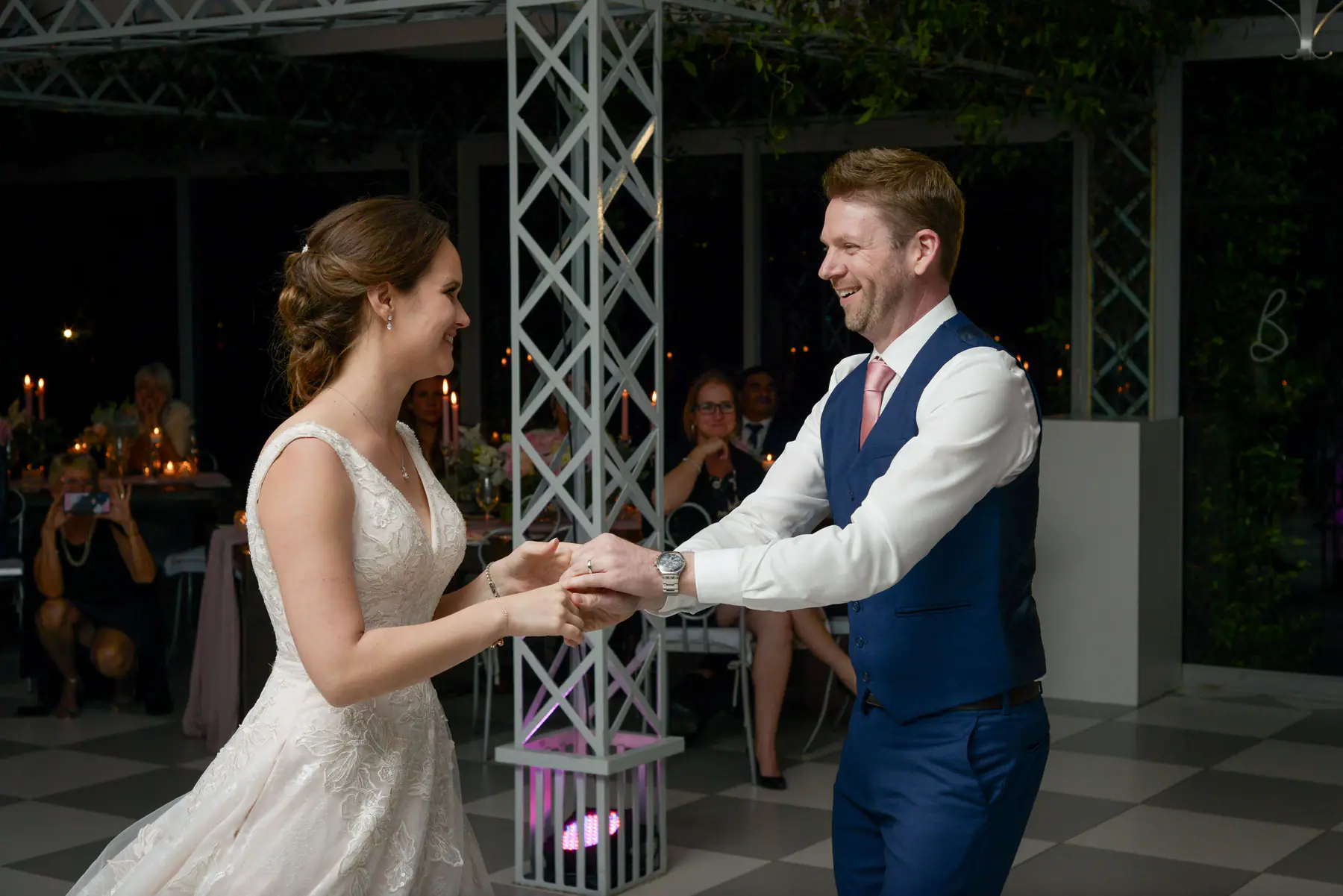 Dora and Scott hand in hand during their first dance as a seated guest films on her phone, a pink uplighter nearby