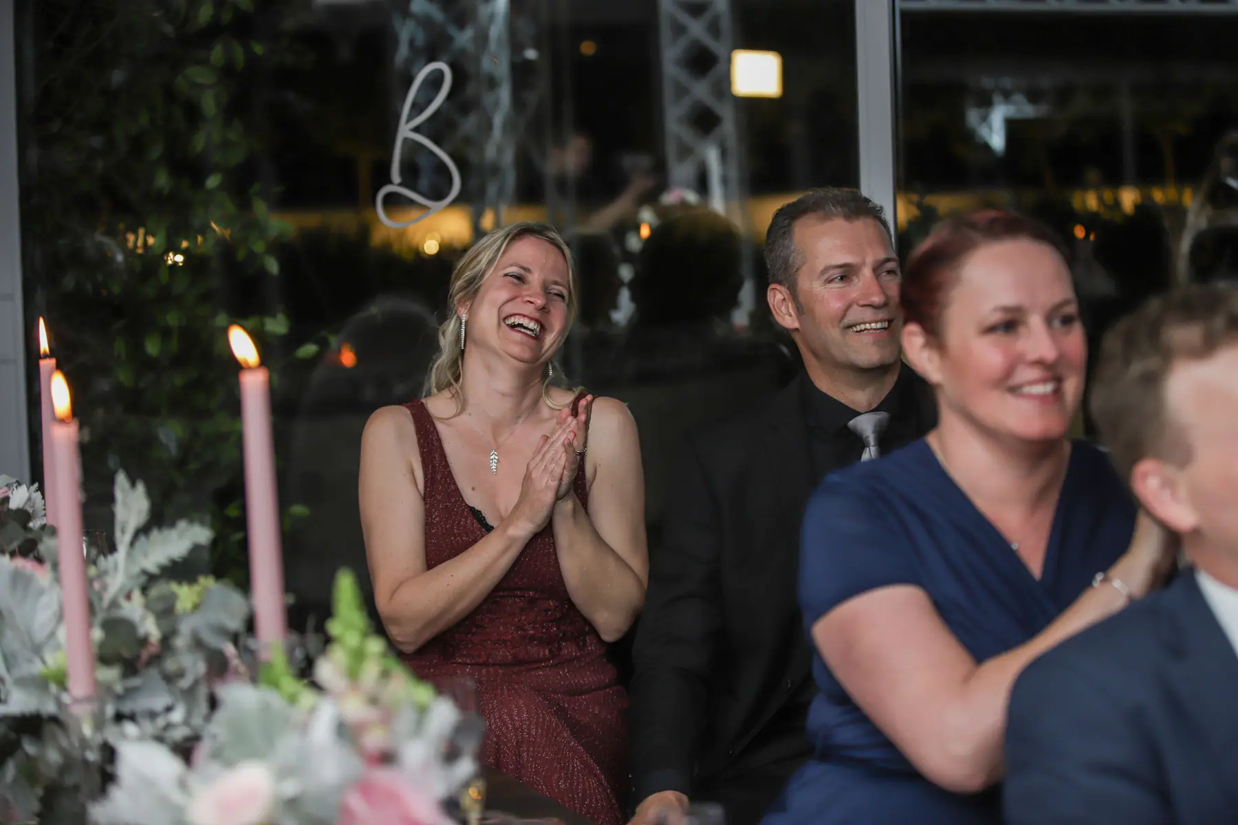 Guests laughing and clapping during the speeches behind pink taper candles, a neon letter B on the window