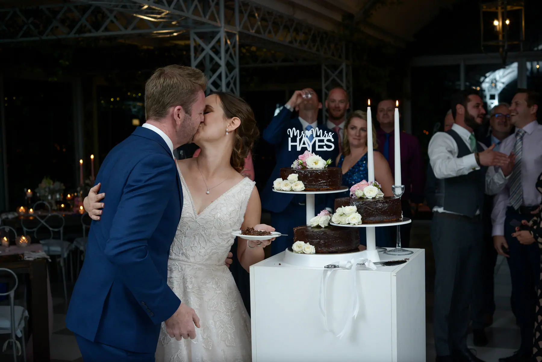 Scott and Dora kissing after cutting their chocolate cake as guests cheer and one downs a shot behind them