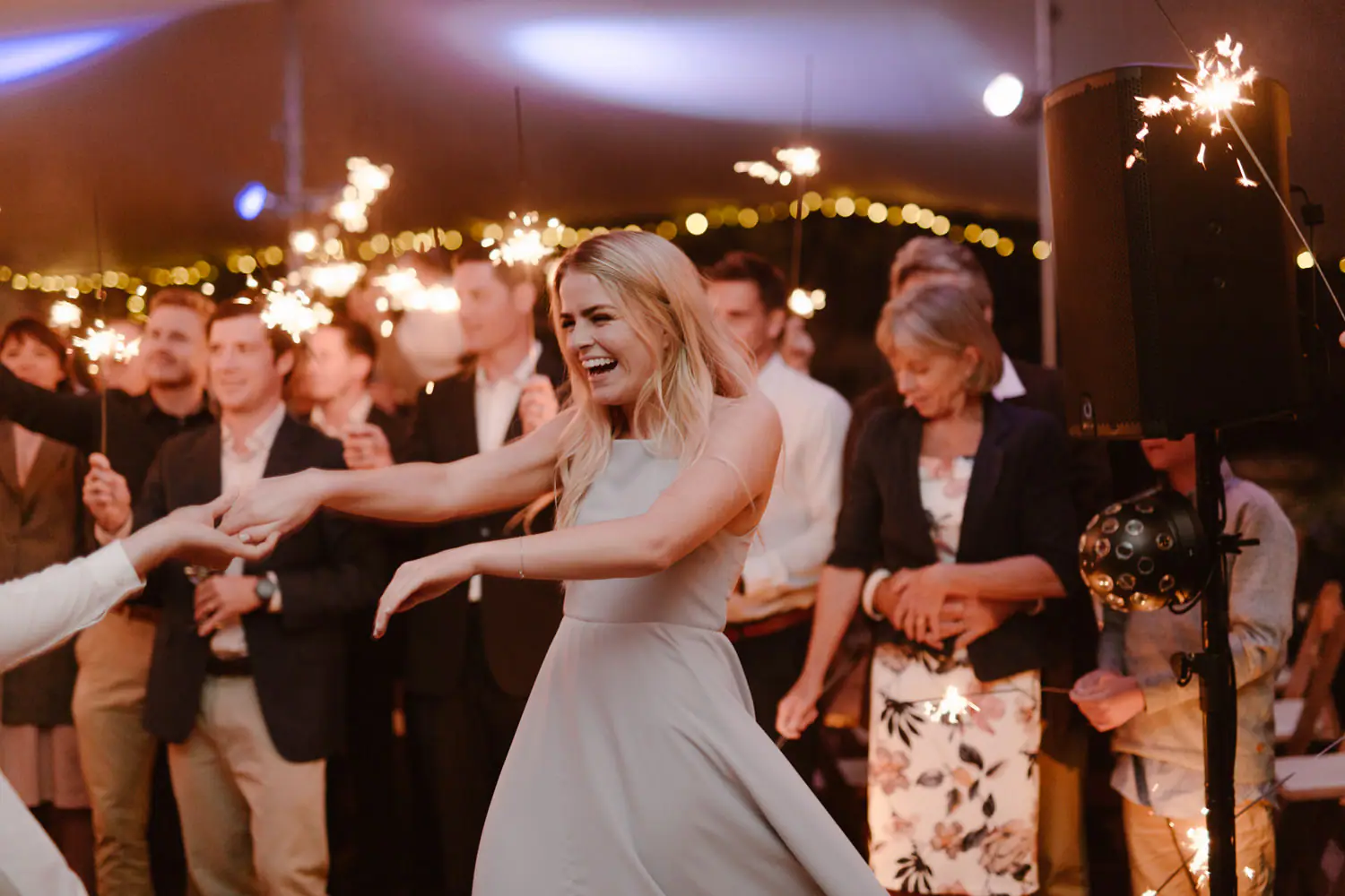 Bridesmaid in a pale grey dress laughing mid-dance as guests hold sparklers, beside a speaker and LED party light on a stand