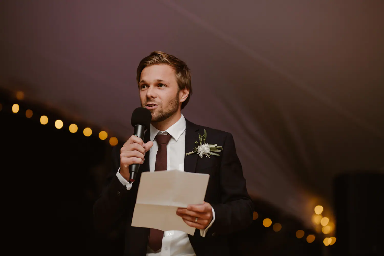 Ramsay reading his groom's speech from a folded page into a microphone, a white flower on his lapel