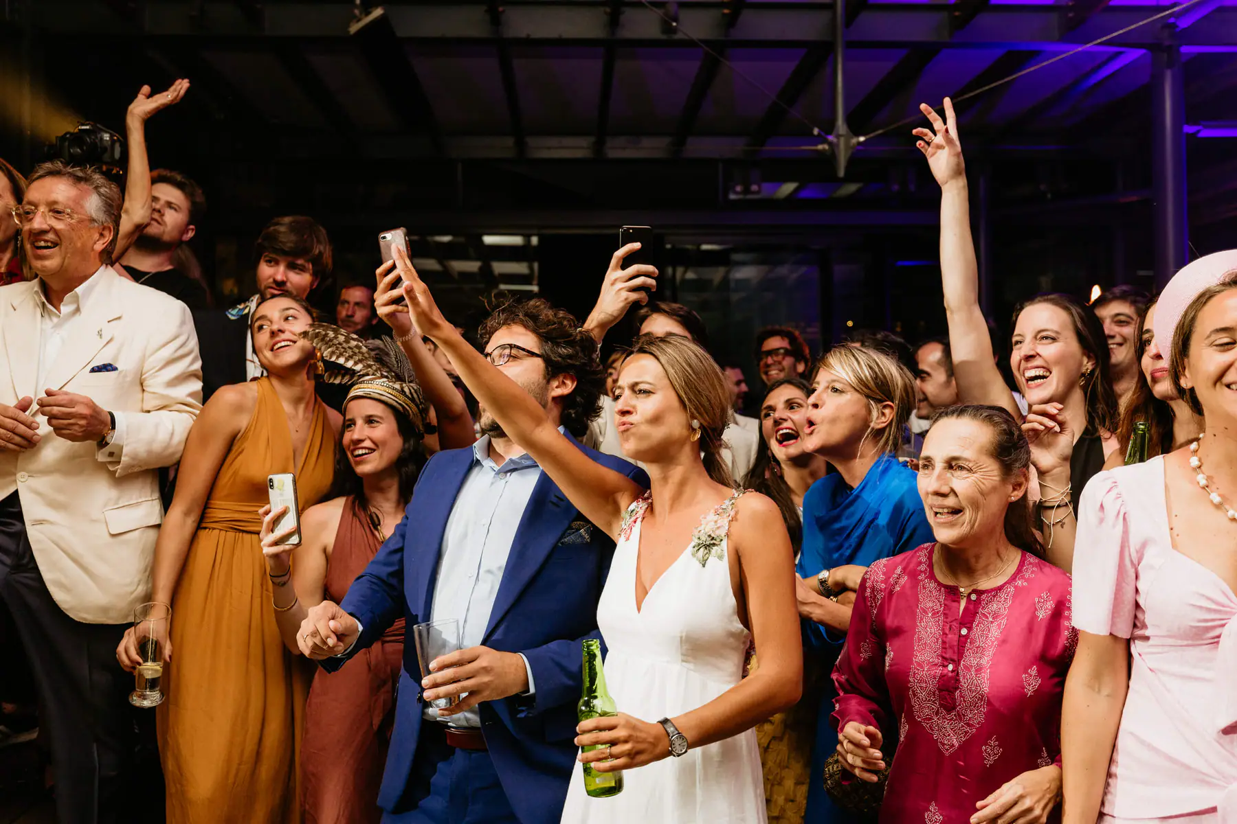 Barbara raises an arm among guests holding phones up to film, Mario beside her with a glass, purple light on the roof