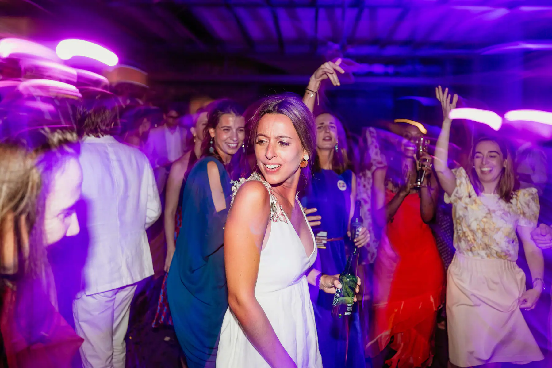 Barbara dances with a beer among friends in blue, red and floral dresses, purple light streaking in motion blur