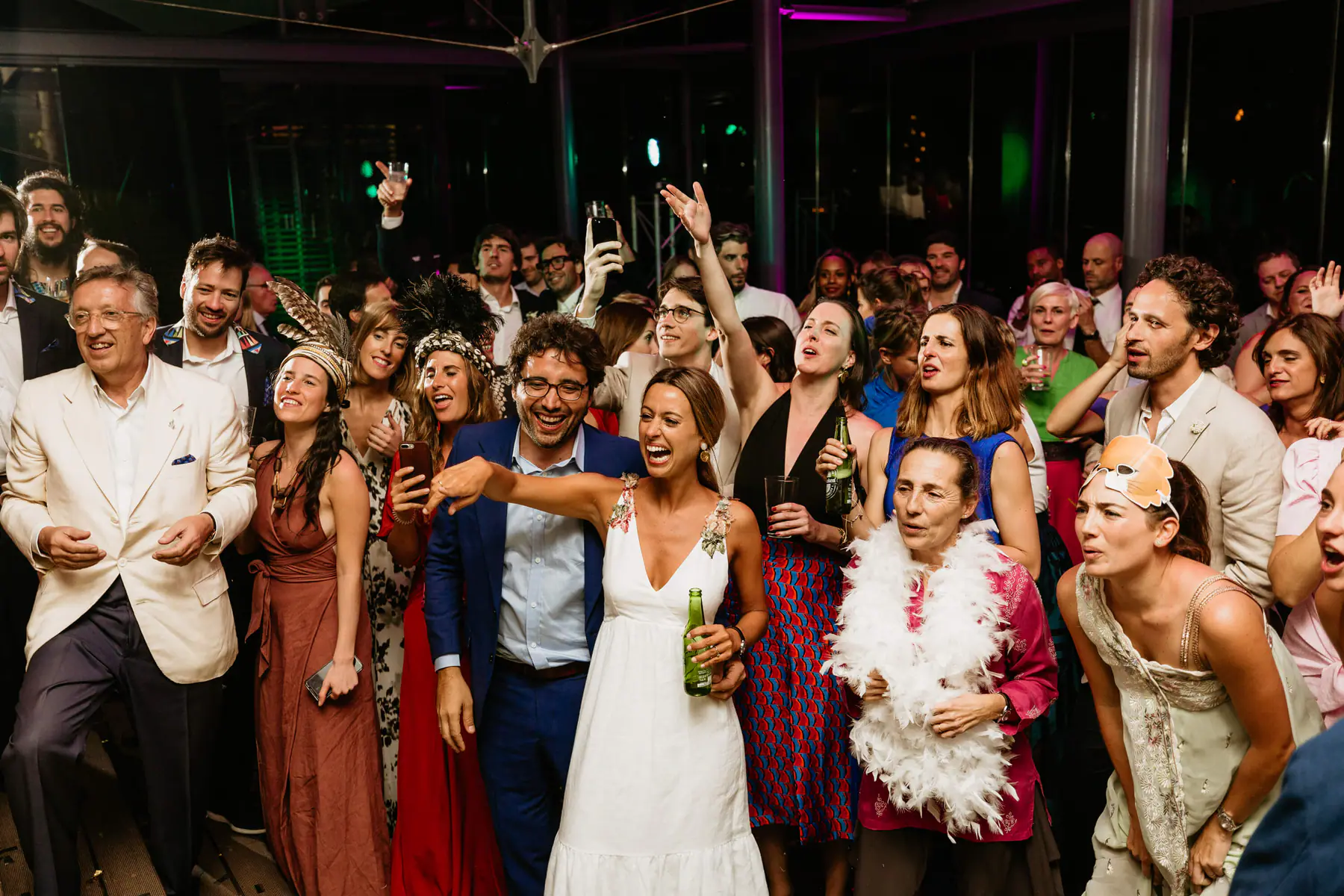 Barbara points and laughs with a beer as Mario grins beside her among guests in feather headdresses