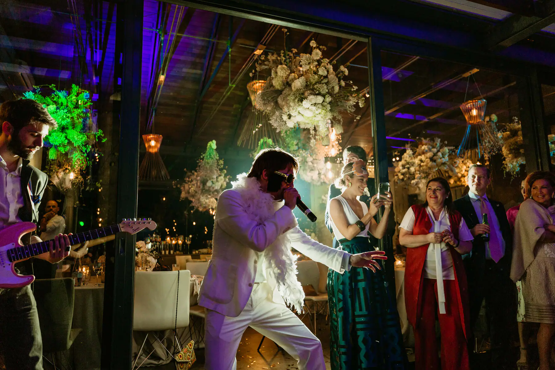 An Elvis impersonator in a white suit sings beside a guitarist with a pink electric guitar under hanging flowers at Tokara