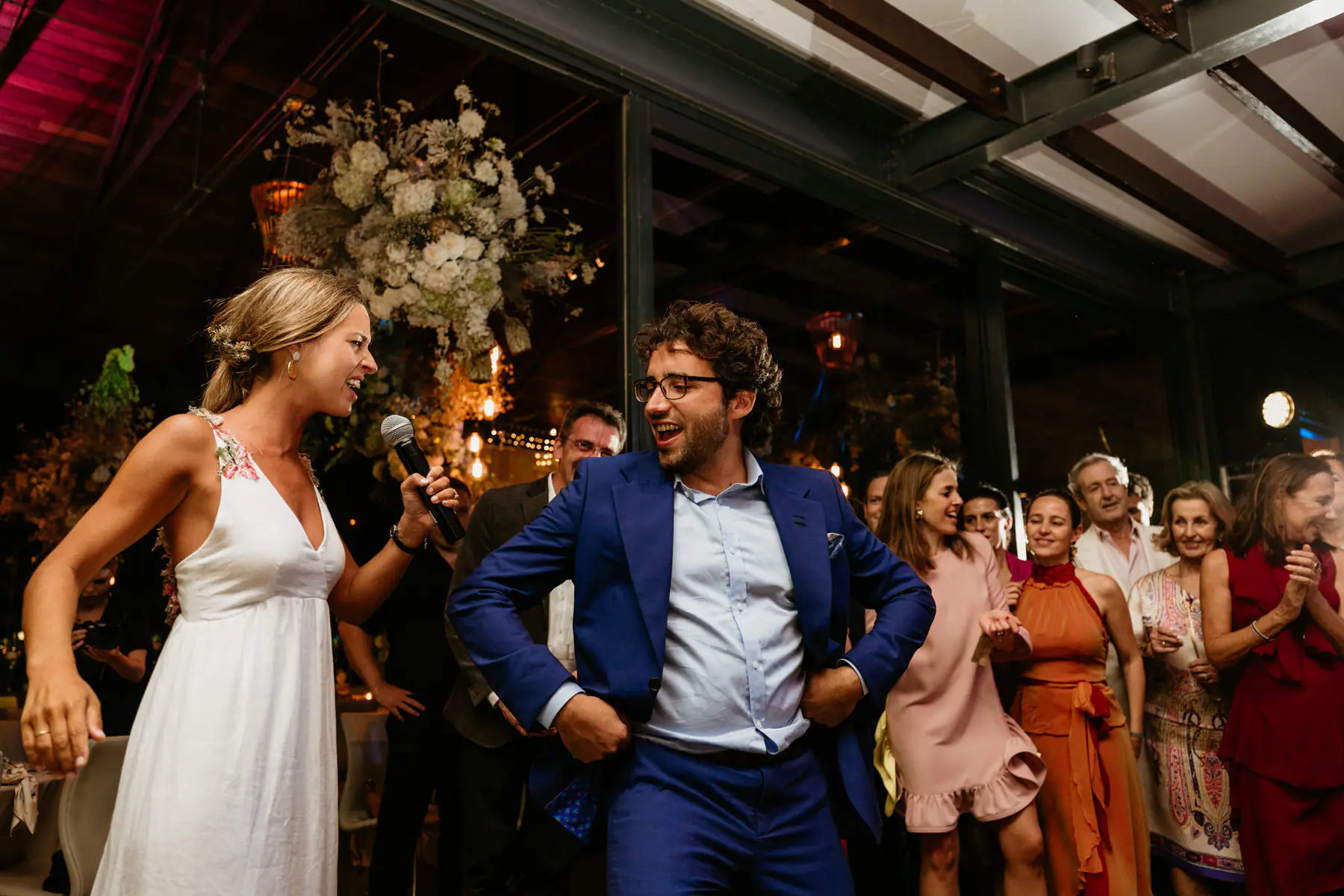 Barbara sings into a microphone as Mario, in glasses and a blue suit, dances for their laughing guests