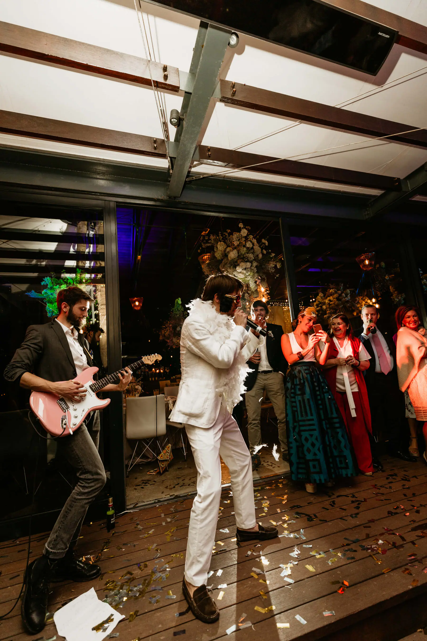 An Elvis impersonator and a guitarist perform on a confetti-strewn wooden deck while guests film them