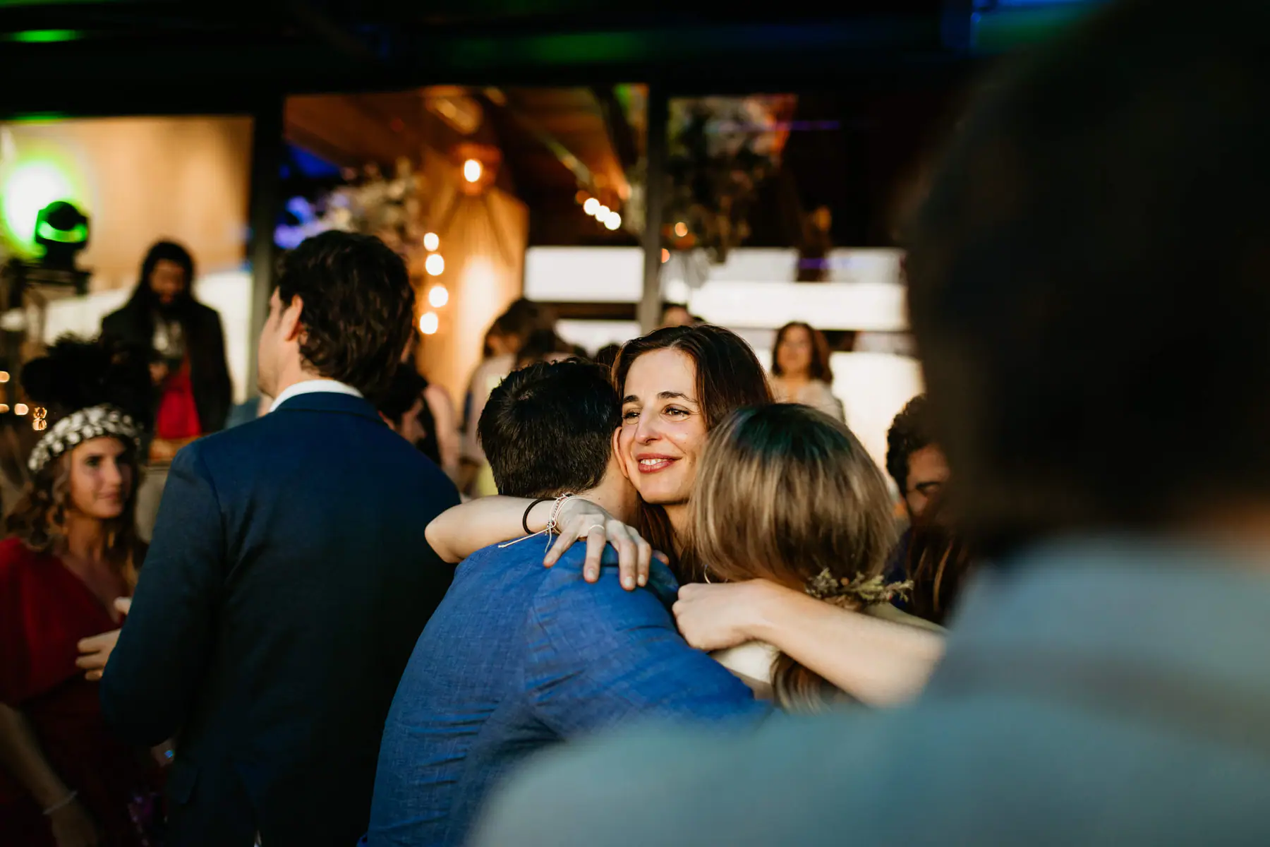 A guest smiles over a group hug on the dance floor, a green-lit moving head light glowing in the background