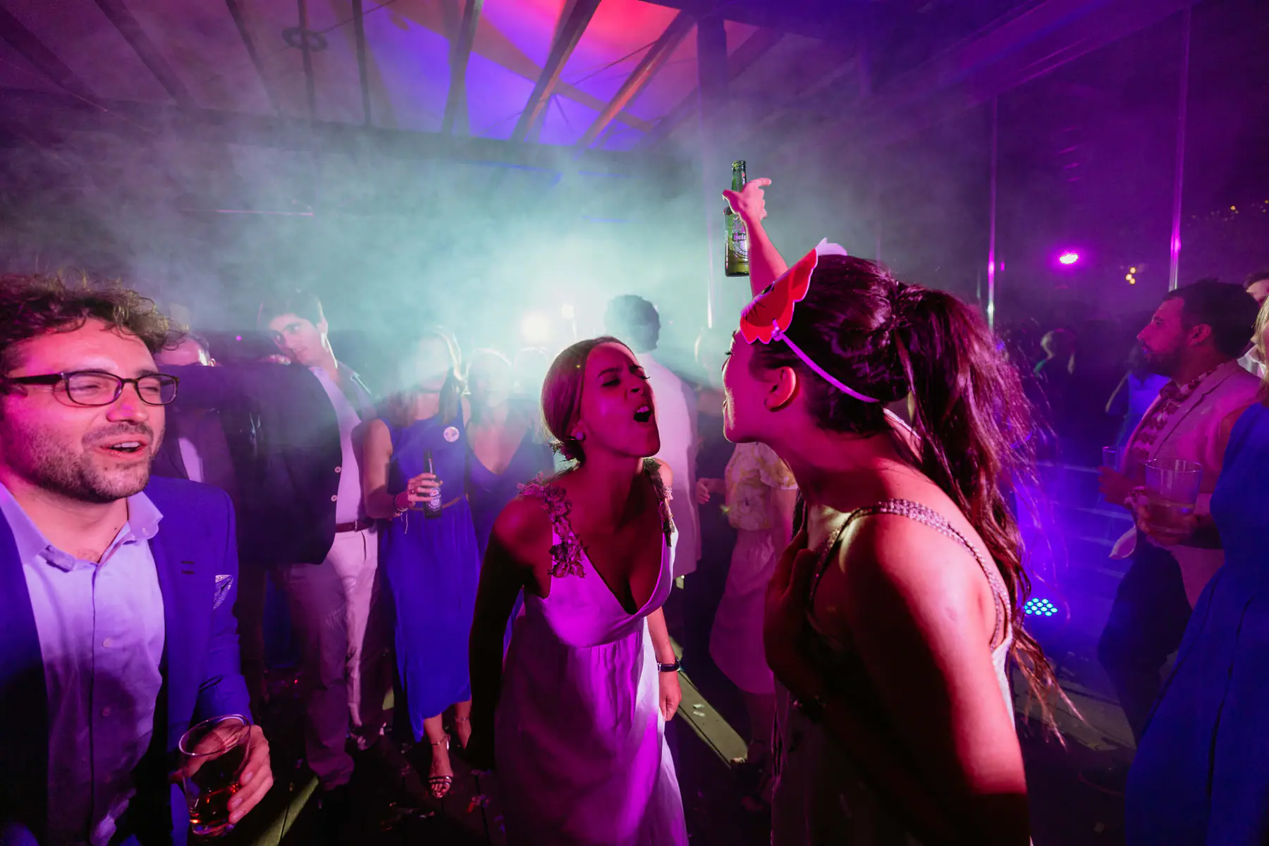 Barbara sings face to face with a friend holding up a beer in pink haze and purple light, Mario laughing beside them