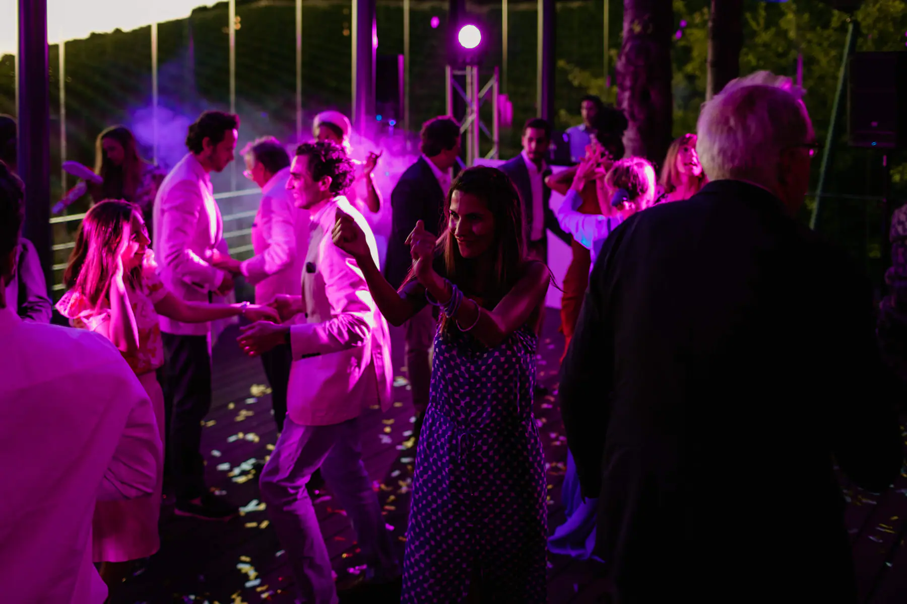 Guests dance on a confetti-scattered deck in magenta light and haze at dusk in Stellenbosch, a truss light behind