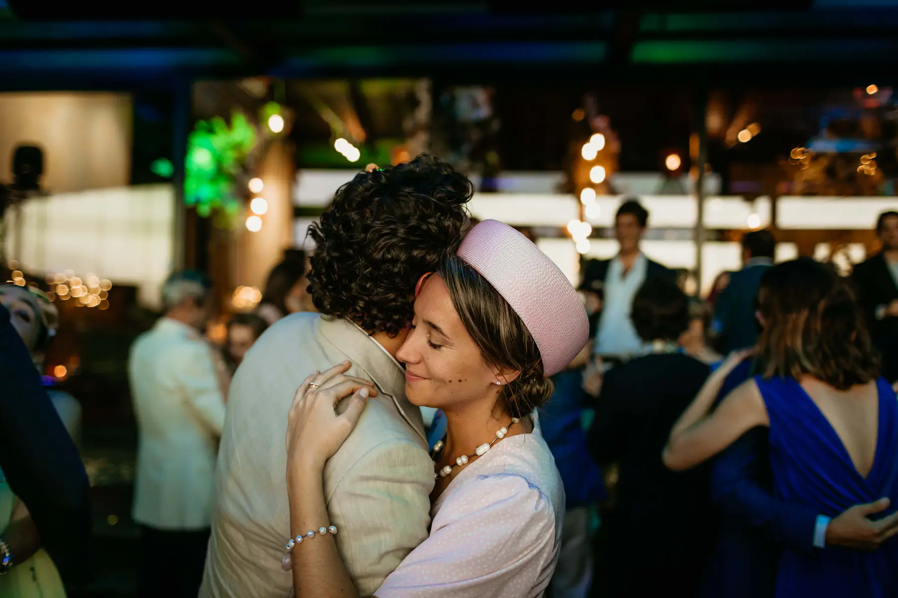 A guest in a pink pillbox hat and pearls slow dances cheek to cheek with a curly-haired man in a linen jacket
