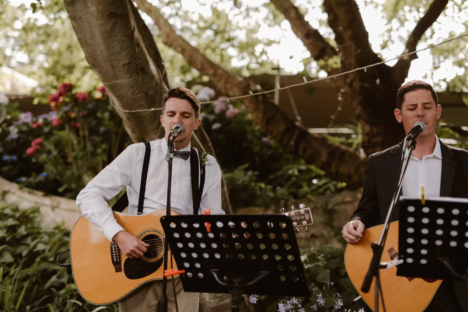 Two musicians singing and playing acoustic guitars behind black music stands in a garden, under a large tree