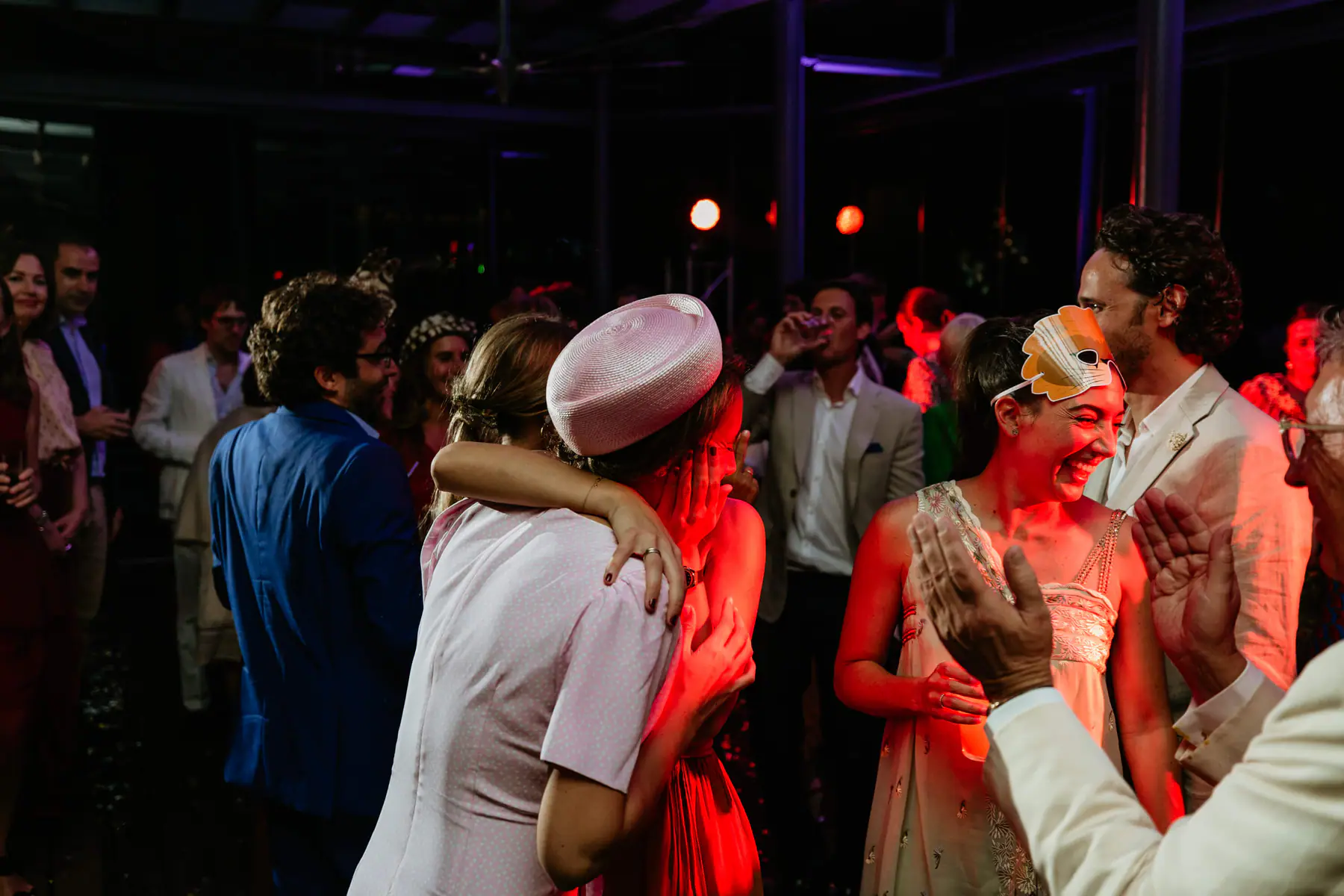 A guest in a pink pillbox hat hugs a friend as a woman with a feathered mask on her head laughs on the red-lit dance floor