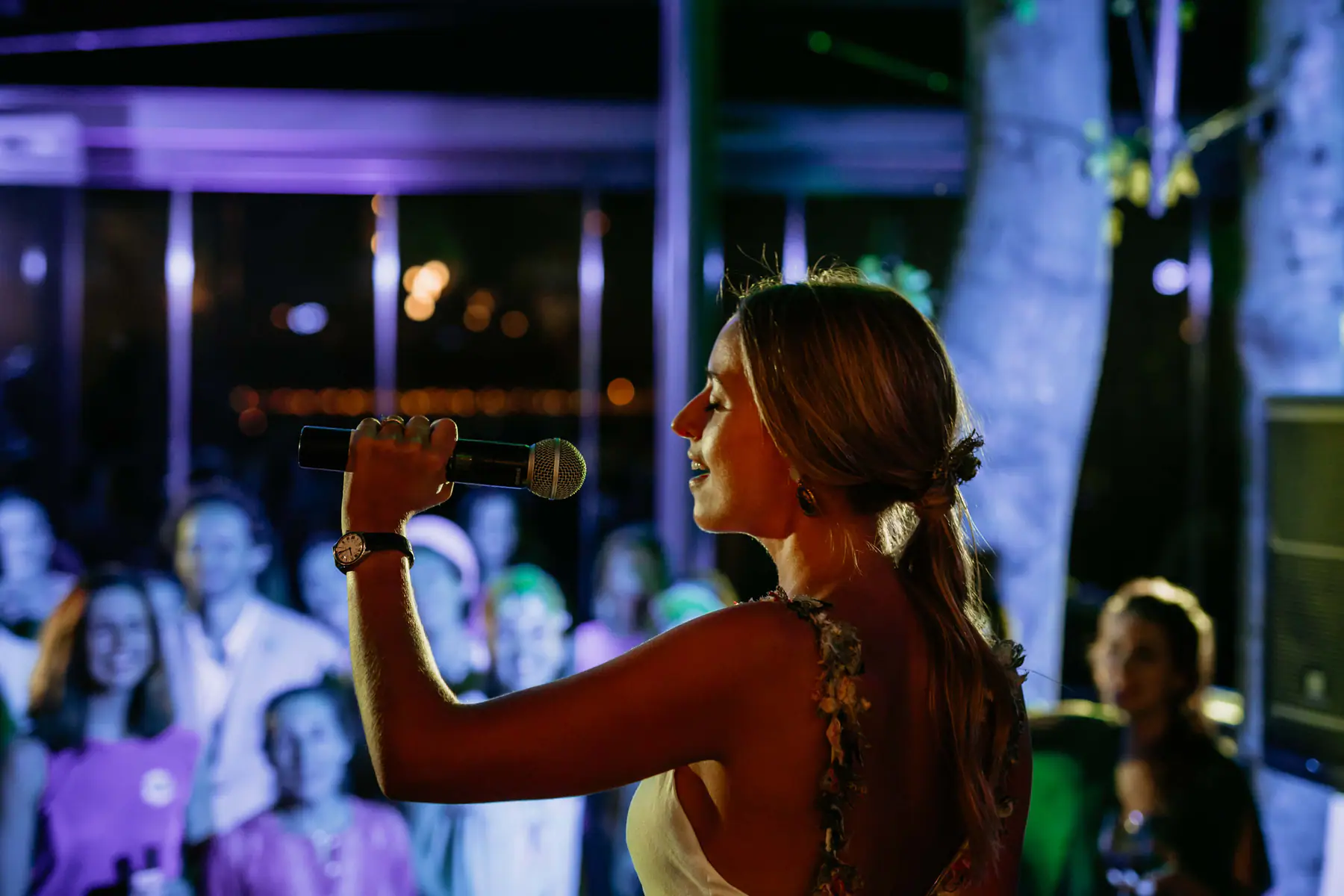 Barbara, in profile, holds up a microphone and smiles at guests lit in purple, a speaker at her side